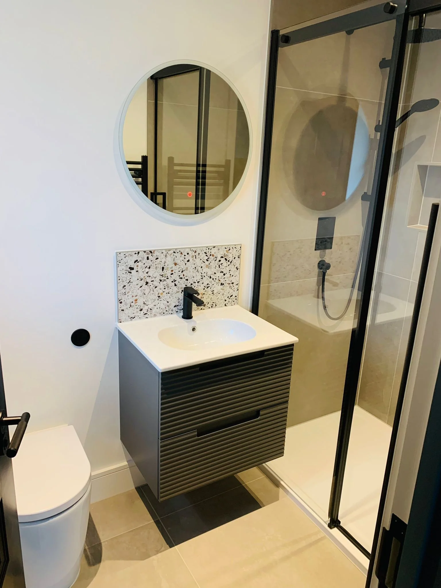 Modern bathroom with a white toilet, a vessel sink with black faucet, a black vanit with slatted drawer design, a terrazzo backsplash, and a walk-in shower with glass door and black fixtures.