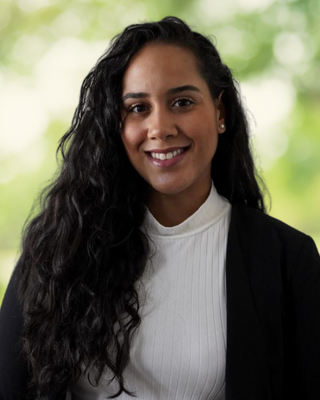 A woman with long, curly black hair smiling, wearing a white shirt and black blazer, standing outdoors with a blurred green background.
