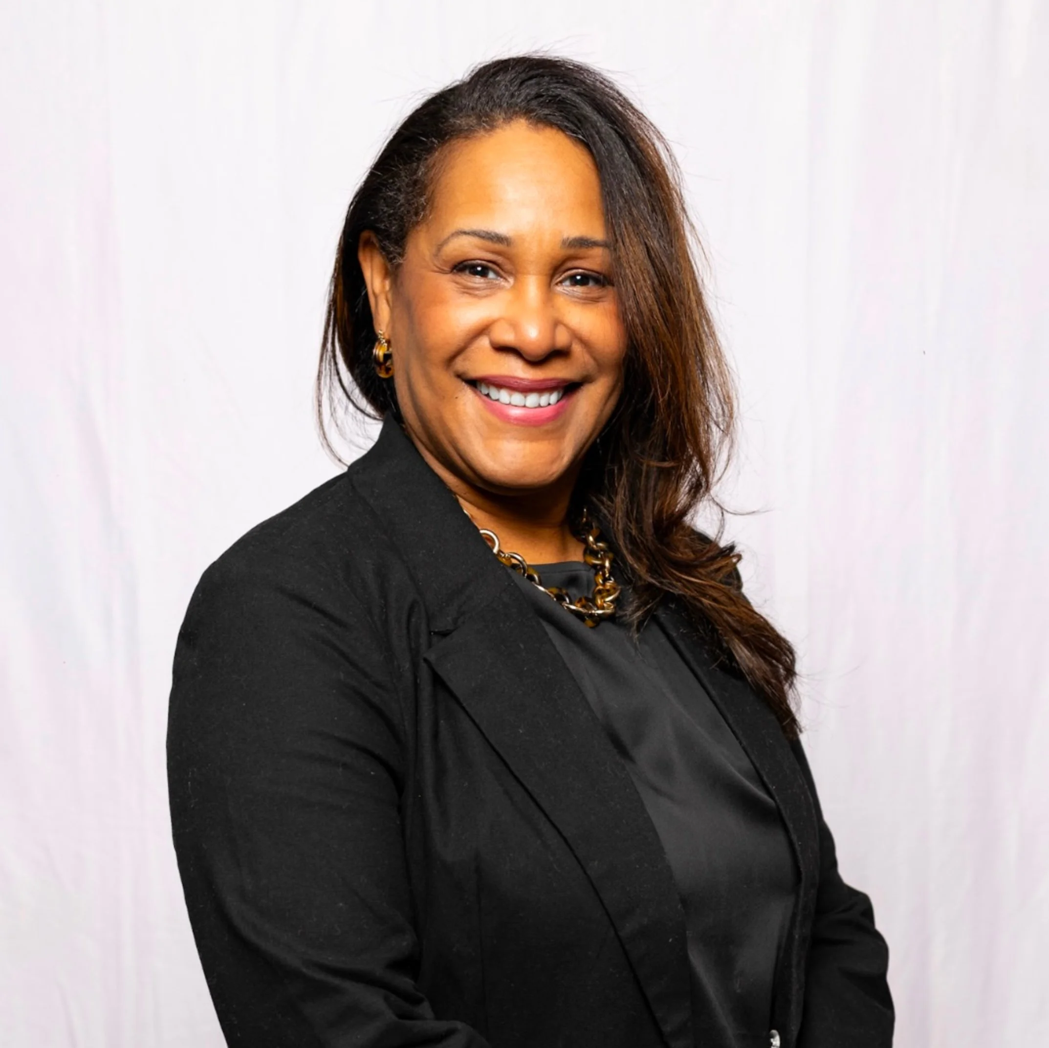 Close-up of a smiling woman with medium skin, wearing gold earrings, a black blazer, and a gold chain necklace, against a plain white background.