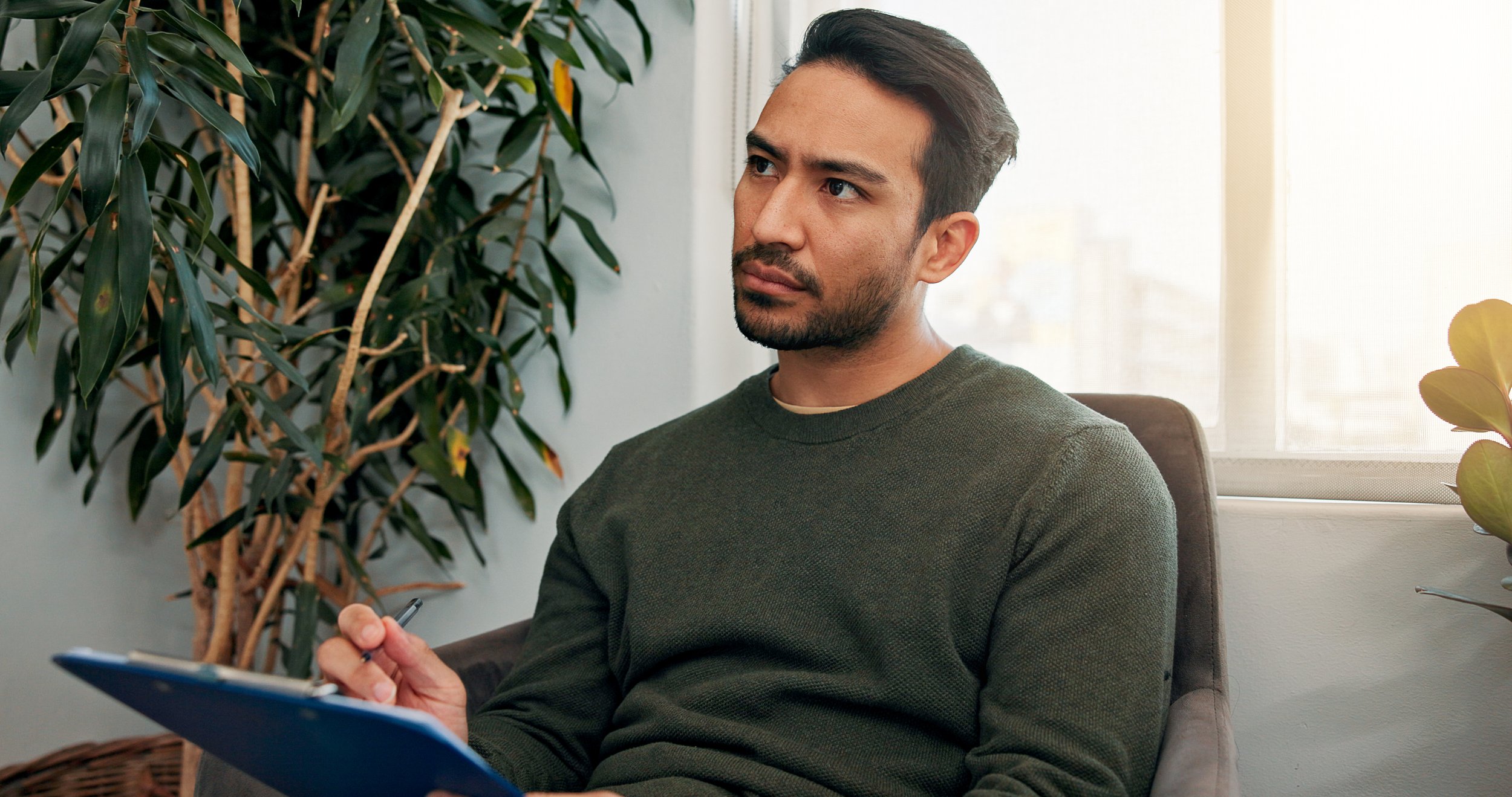 A man with dark hair and light skin is sitting by a window, holding a ceramic mug in his right hand, and looking thoughtfully outside. He is wearing a dark green button-up shirt, and the background shows an industrial-style interior with wooden and metal elements.