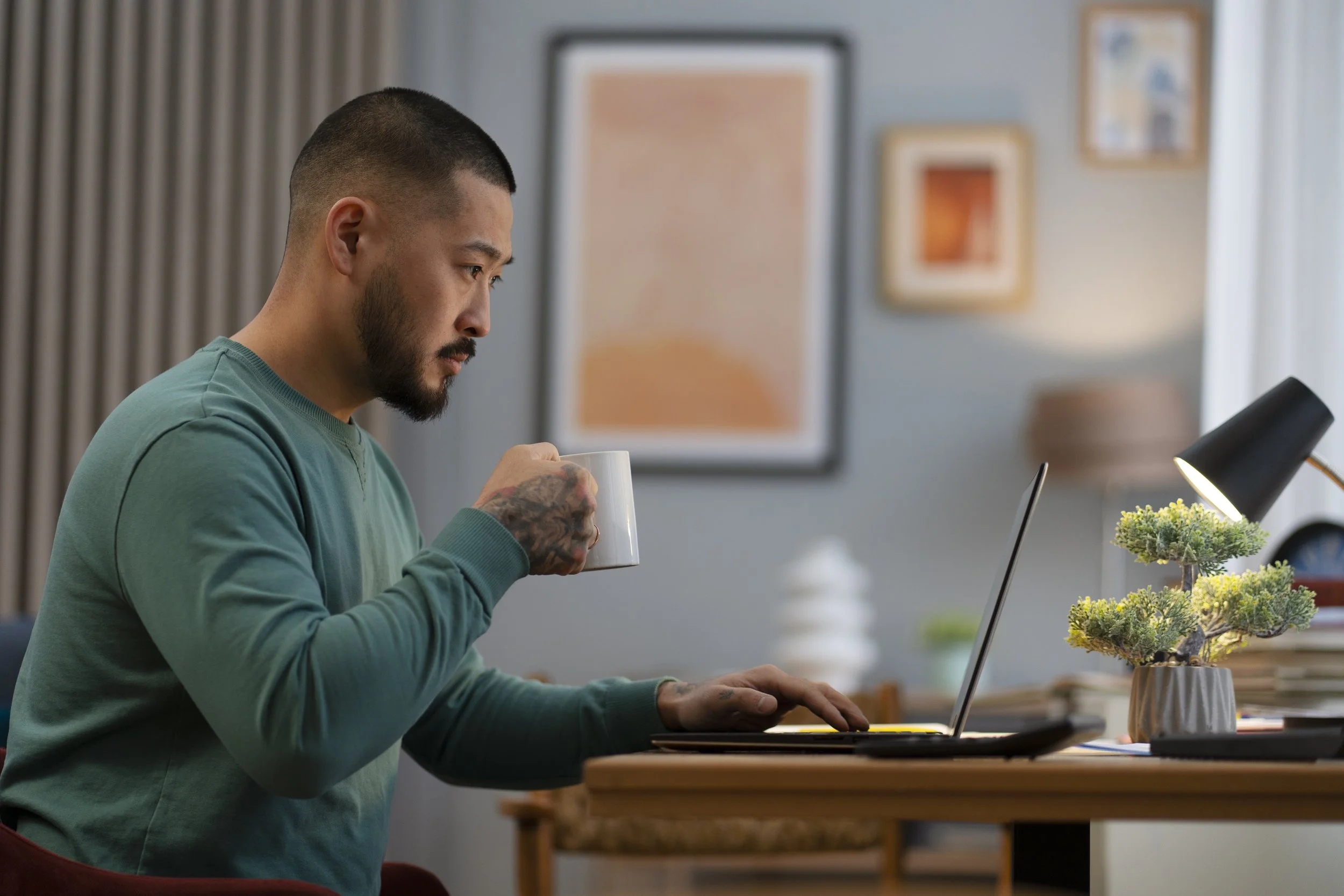 A man with a beard and tattoos wearing a green sweatshirt sits at a table in a modern, brightly lit room, drinking from a white mug while working on a laptop.