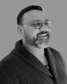 Black and white portrait of a smiling man with glasses, a beard, and short hair, wearing a textured sweater.