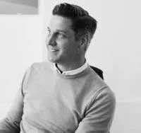 A man smiling, wearing a light sweater and collared shirt, sitting and looking to his right in black and white.