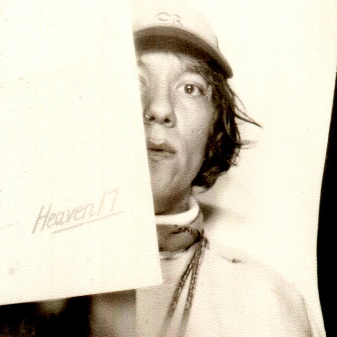 Black and white photo of a young person with curly hair, wearing a cap, holding up the front cover of Heaven 17's 'We Don't Need That Fascist Groove Thing', partially covering their face.