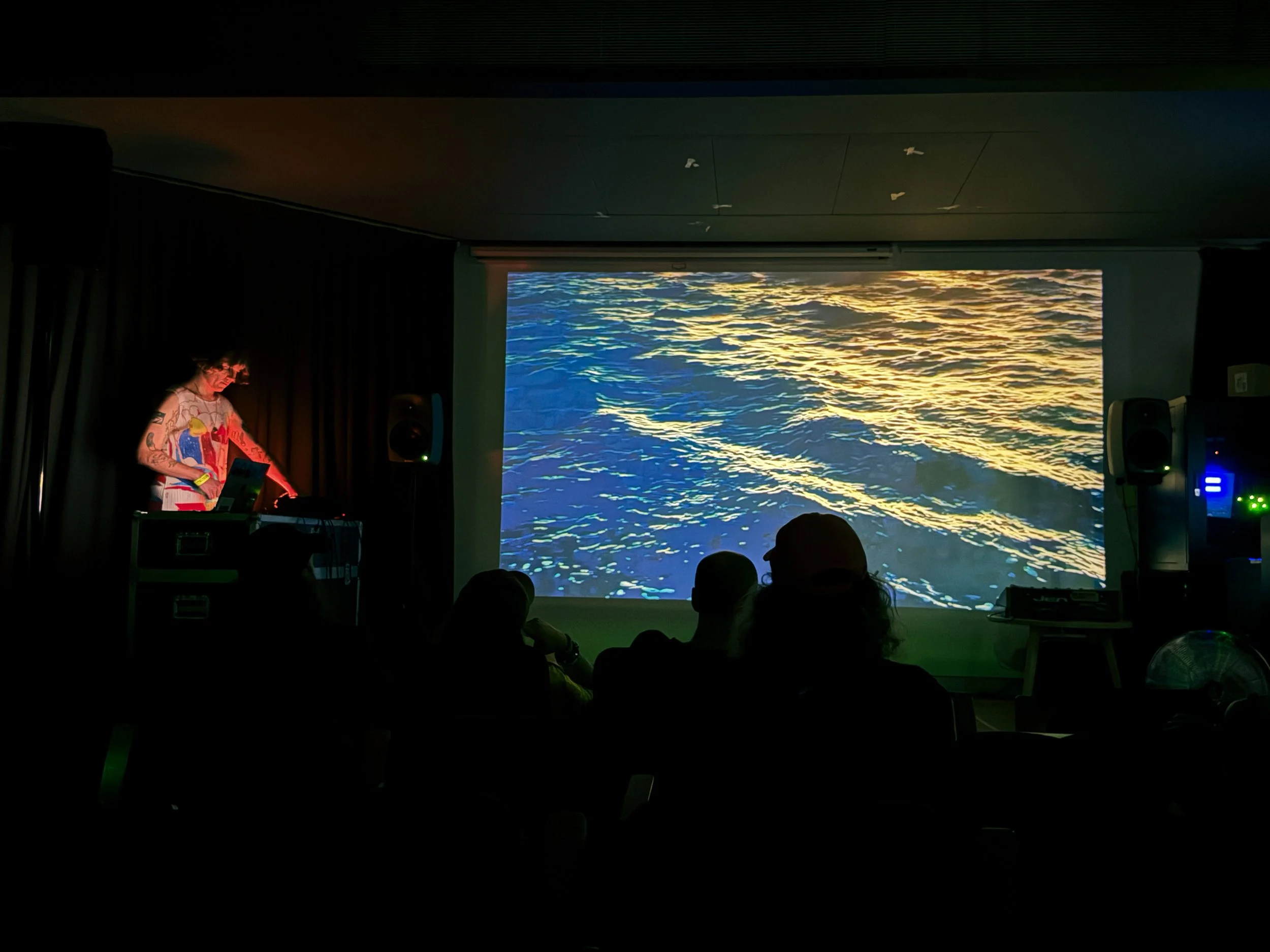 A person playing to the side of the stage using electronic equipment. The majority of the stage is taken up by a huge projection of sea waves.