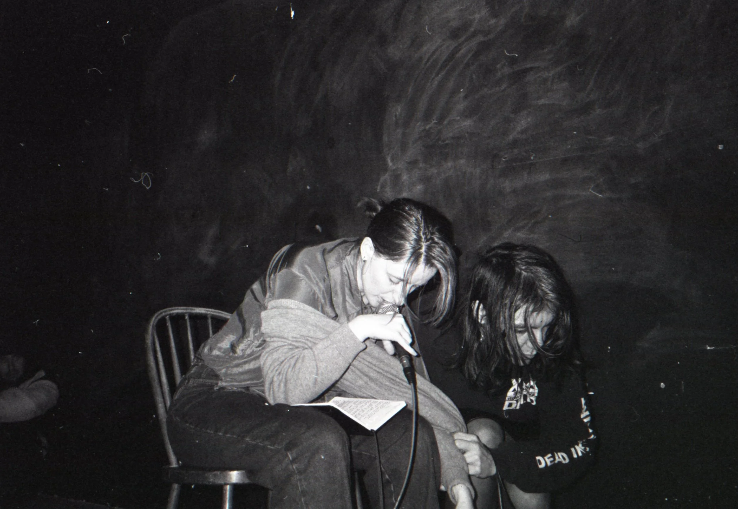 Two people with long dark hair sitting closely together in a dark room. One is pulling at the other's clothing, while the other holds a microphone close to their face with their eyes closed. A notebook is open in their lap.
