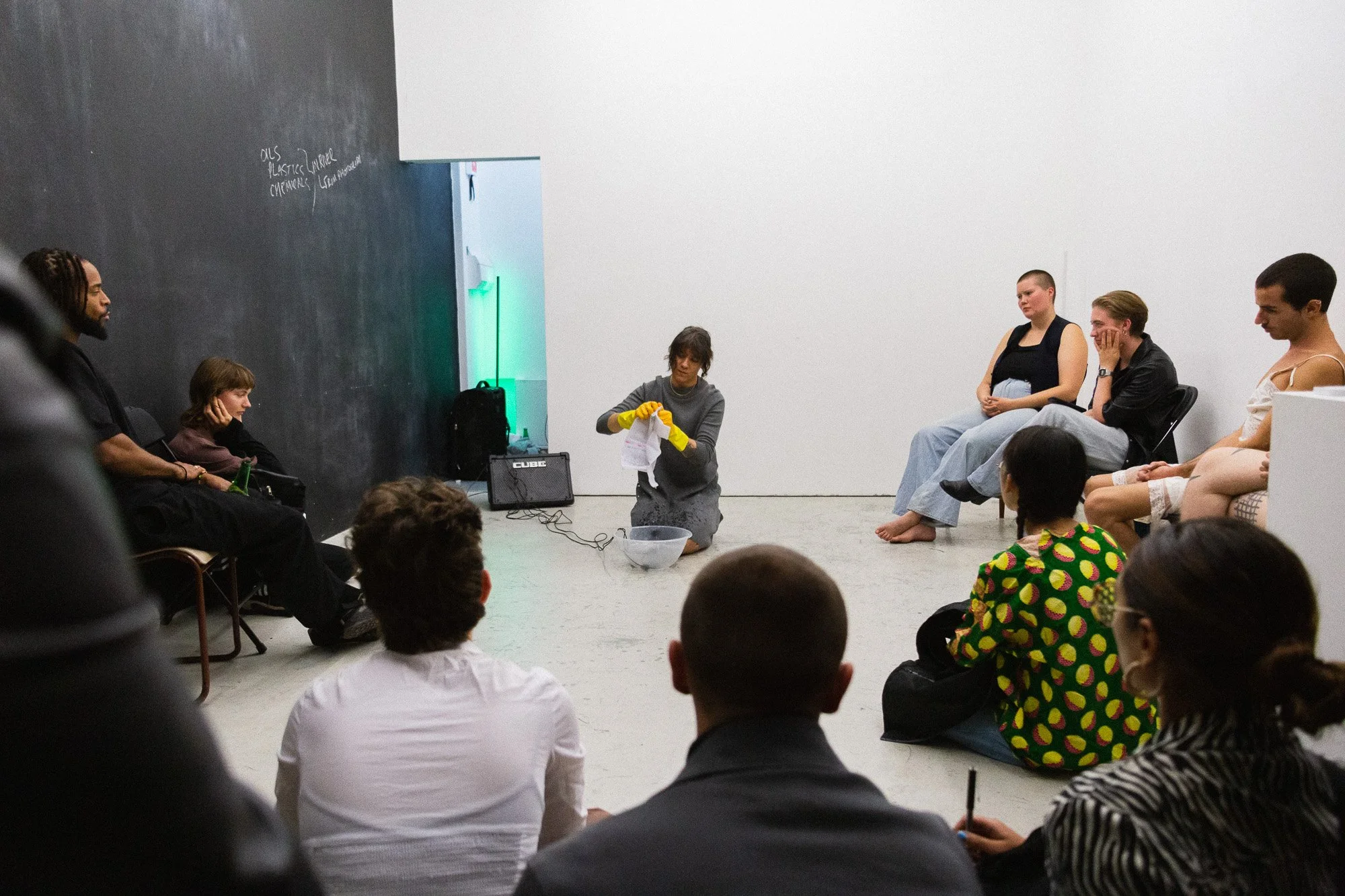 A woman kneeling on the floor with yellow gloves washing or scrubbing an object while a group of people sit around her, observing. The setting appears to be an indoor space with white and black walls. There are wires coming from the washing gloves.