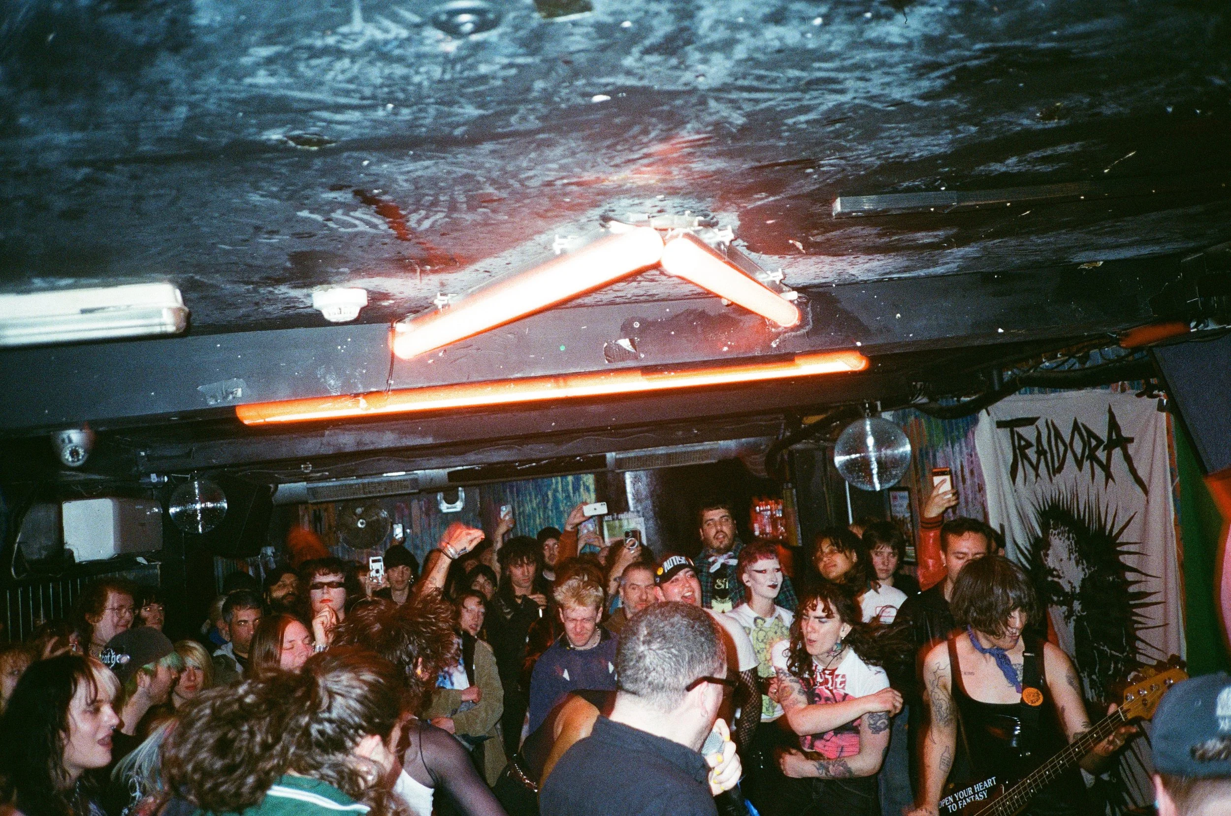 A crowd of people pictured in mid-movement gathered in a dimly lit venue with dark walls, hanging light fixtures, and intense-looking banners while a band plays.