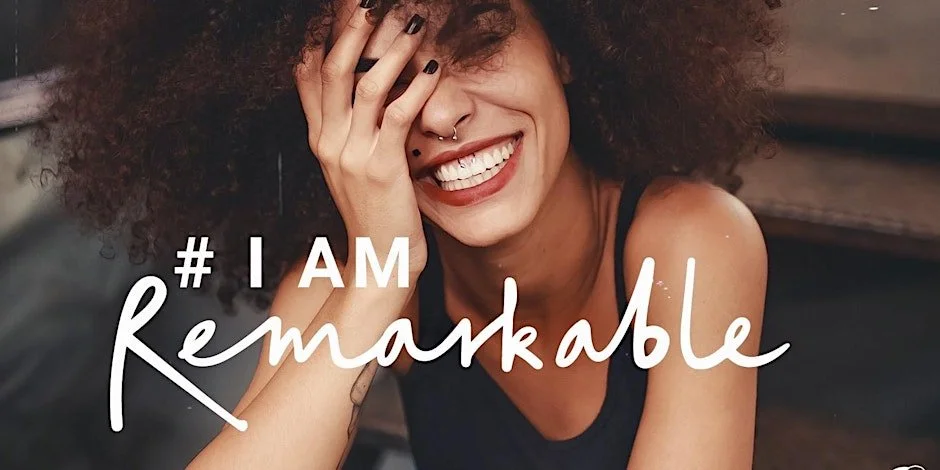 A woman with curly hair and a big smile covering part of her face with her hand, wearing a black tank top, with the text '# I AMremarkable' overlaid.