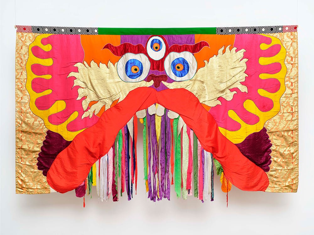 Colorful textile art piece resembling a dragon with prominent eyes, large red tongue, and intricate textures and patterns.