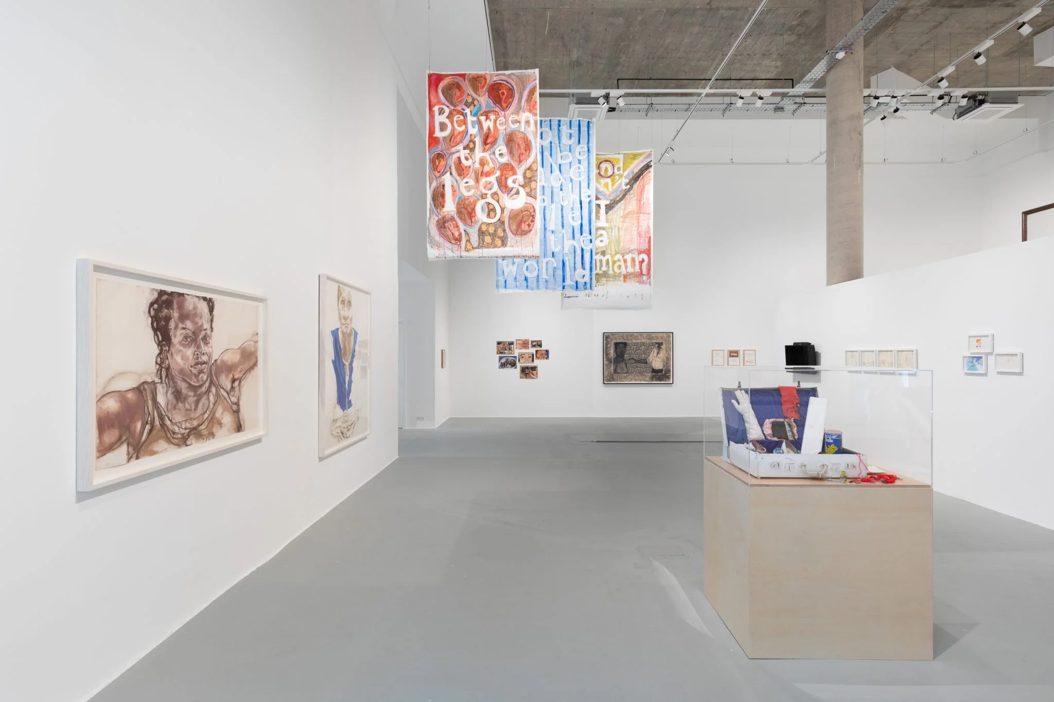 Interior of an art gallery with white walls displaying various paintings. Two hanging textile art pieces with text are visible, one says 'Between the tall trees' and the other features geometric patterns. Several smaller artworks are on the wall in the background. There is a light-colored wooden table with art supplies underneath, and the ceiling has industrial-style exposed pipes and track lighting.