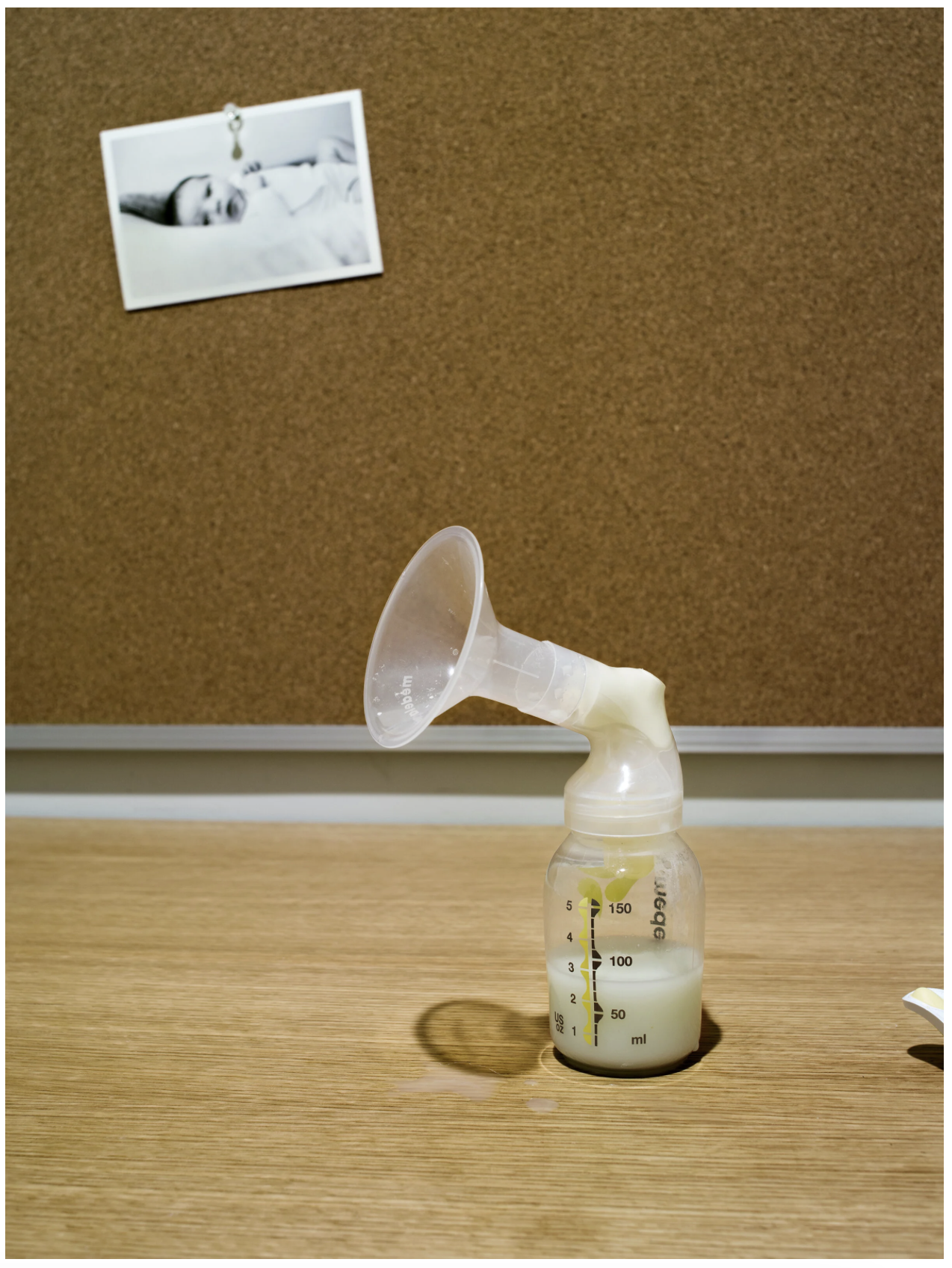 A breastpump with a yellow container and a transparent flange on a wooden surface, with a corkboard and a black-and-white photo of a sleeping baby in the background.
