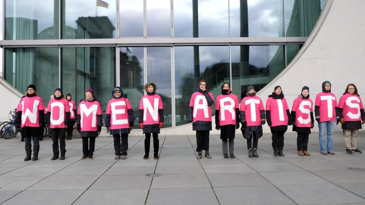 Group of women standing outside modern building, each wearing a pink shirt with a large white letter on front spelling out 'WOMEN ARTISTS'.