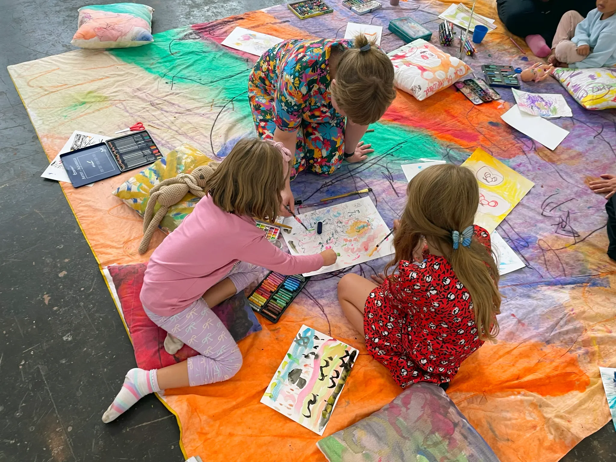 Kids gathered on a colorful fabric spread on the floor, engaging in painting and drawing activities with various art supplies, including markers, watercolors, and paper.