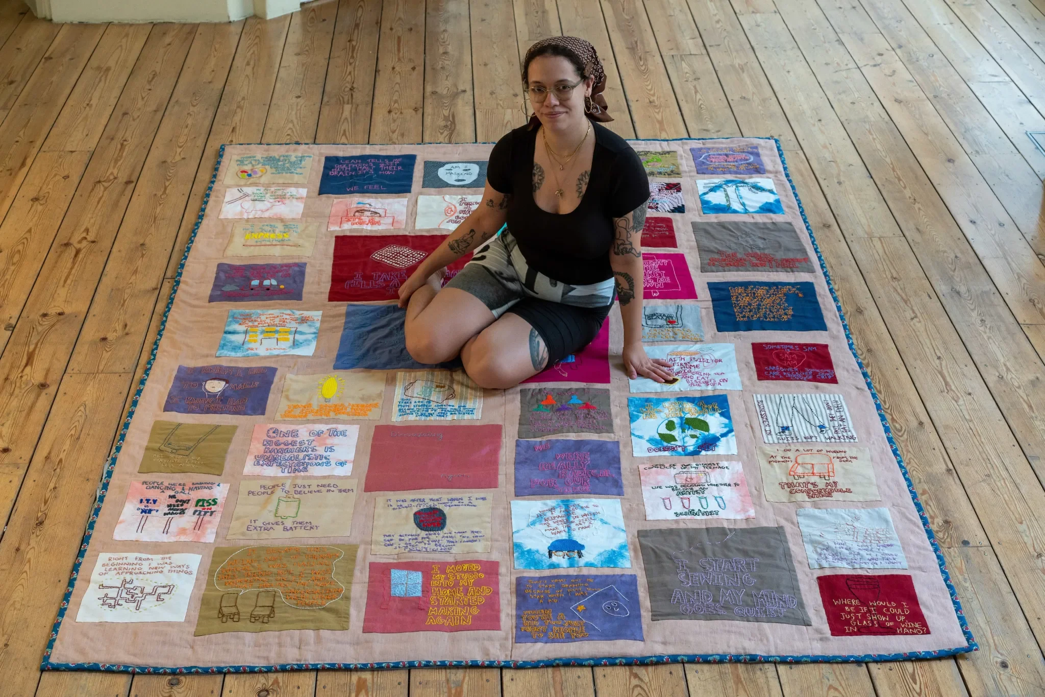 A woman sitting on a colorful quilt with embroidered patches on a wooden floor near a white door.