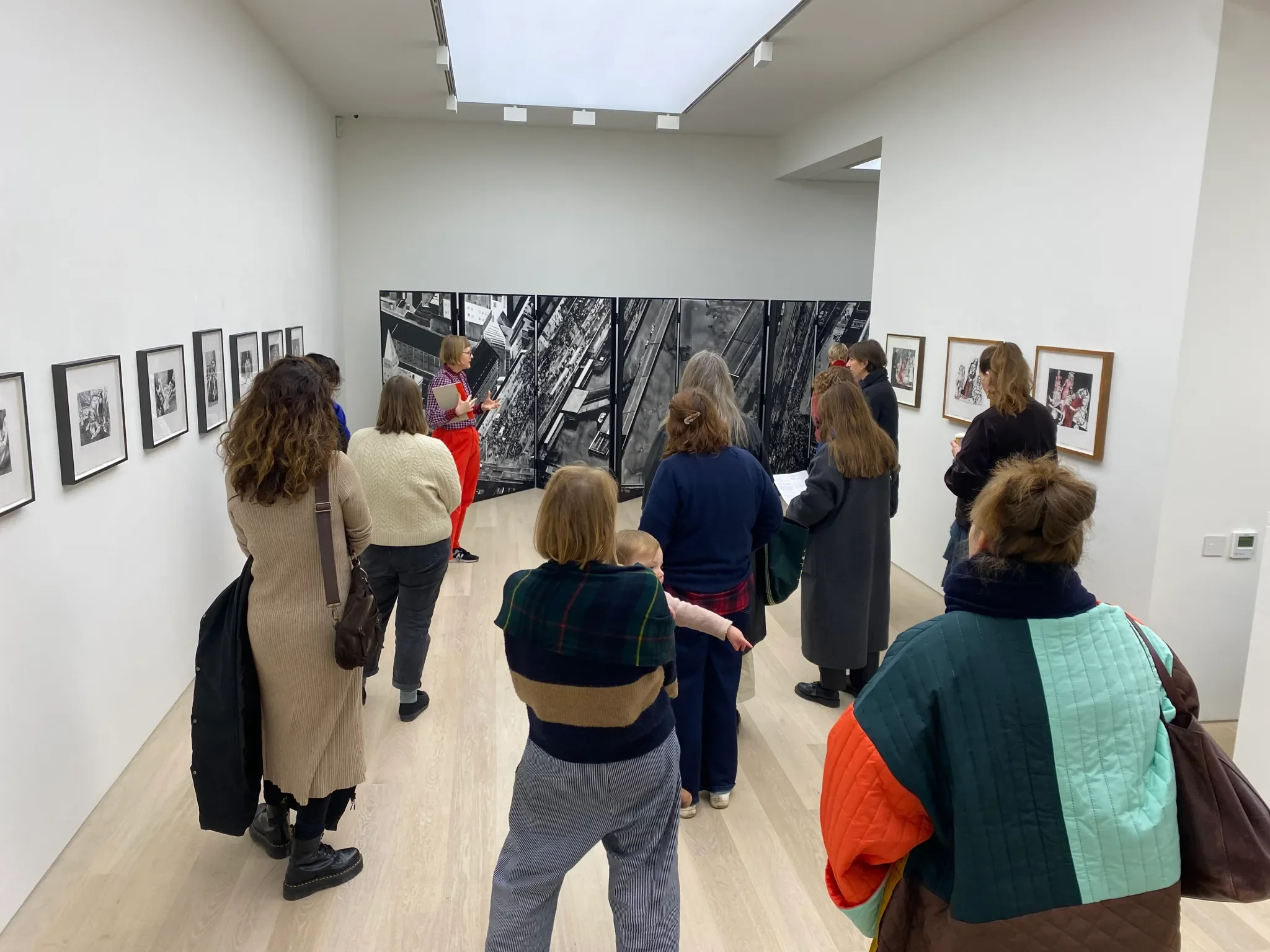 A group of people gathered in an art gallery, listening to a woman in red pants and a checkered shirt explaining artwork. The gallery has framed art on the walls and large black-and-white photographs on display partitions.