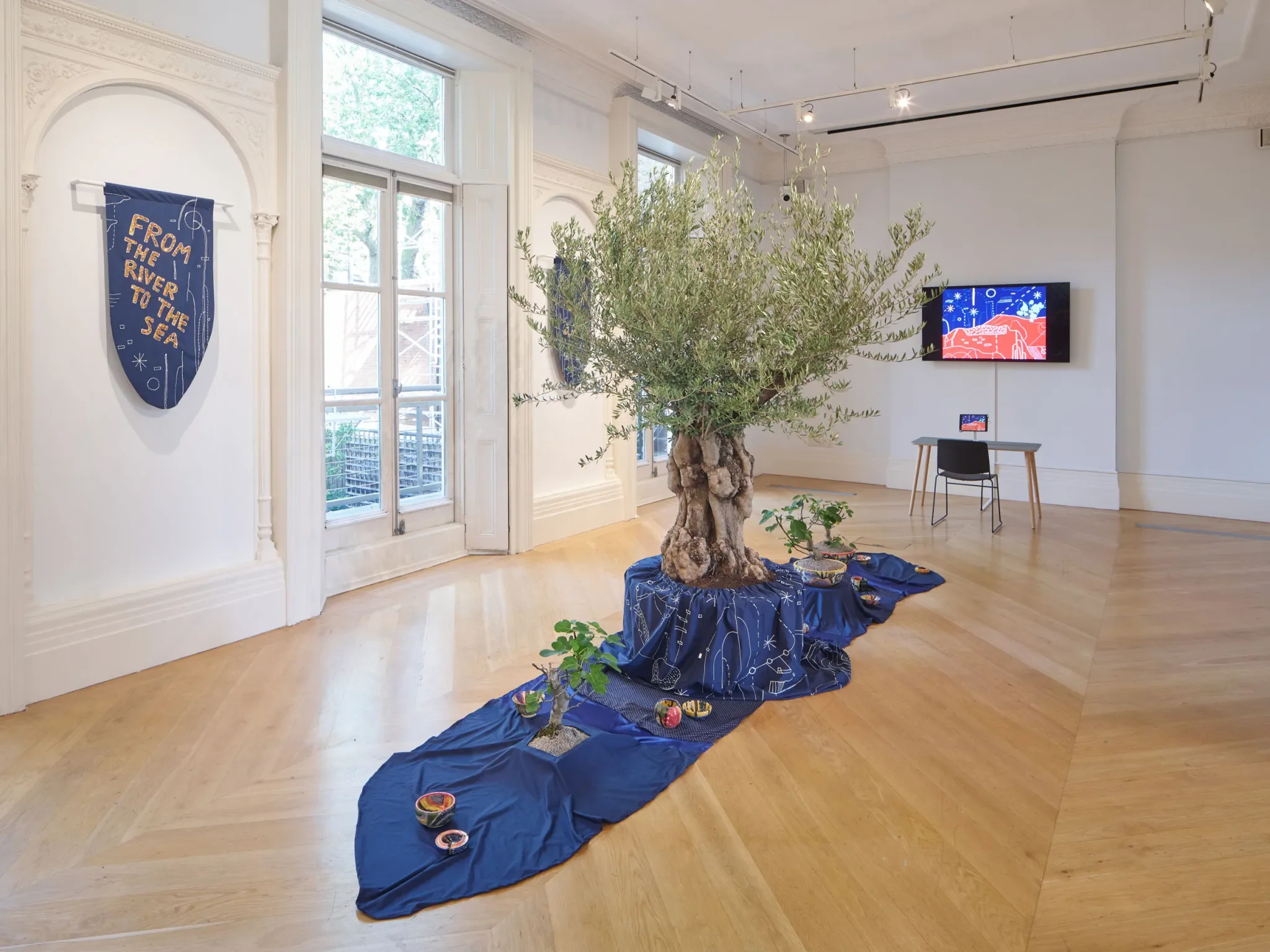 Museum or gallery interior with a large olive tree in the center, surrounded by a blue fabric cloth with white constellations, and small plant pots. On the wall, there is a blue banner with the text 'From the River to the Sea'. A television screen displays a colorful digital illustration. Large windows provide natural light.
