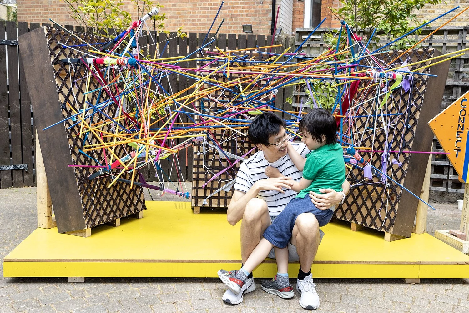 A man and a young boy smiling and playing on a yellow platform in front of a colorful string art display outdoors. The man is sitting on the platform, holding the boy, who is touching his face.