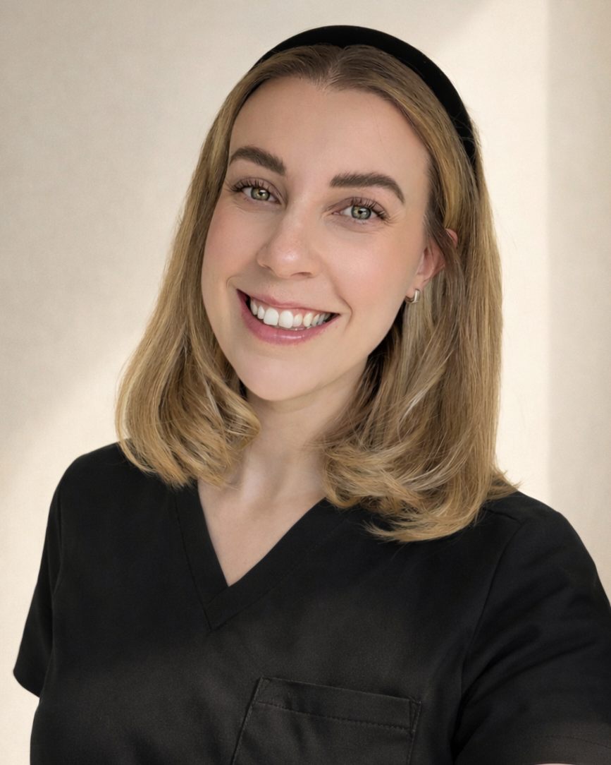 A young woman with blonde hair, wearing black scrubs and a headband, smiling at the camera. The background is plain and neutral-colored.