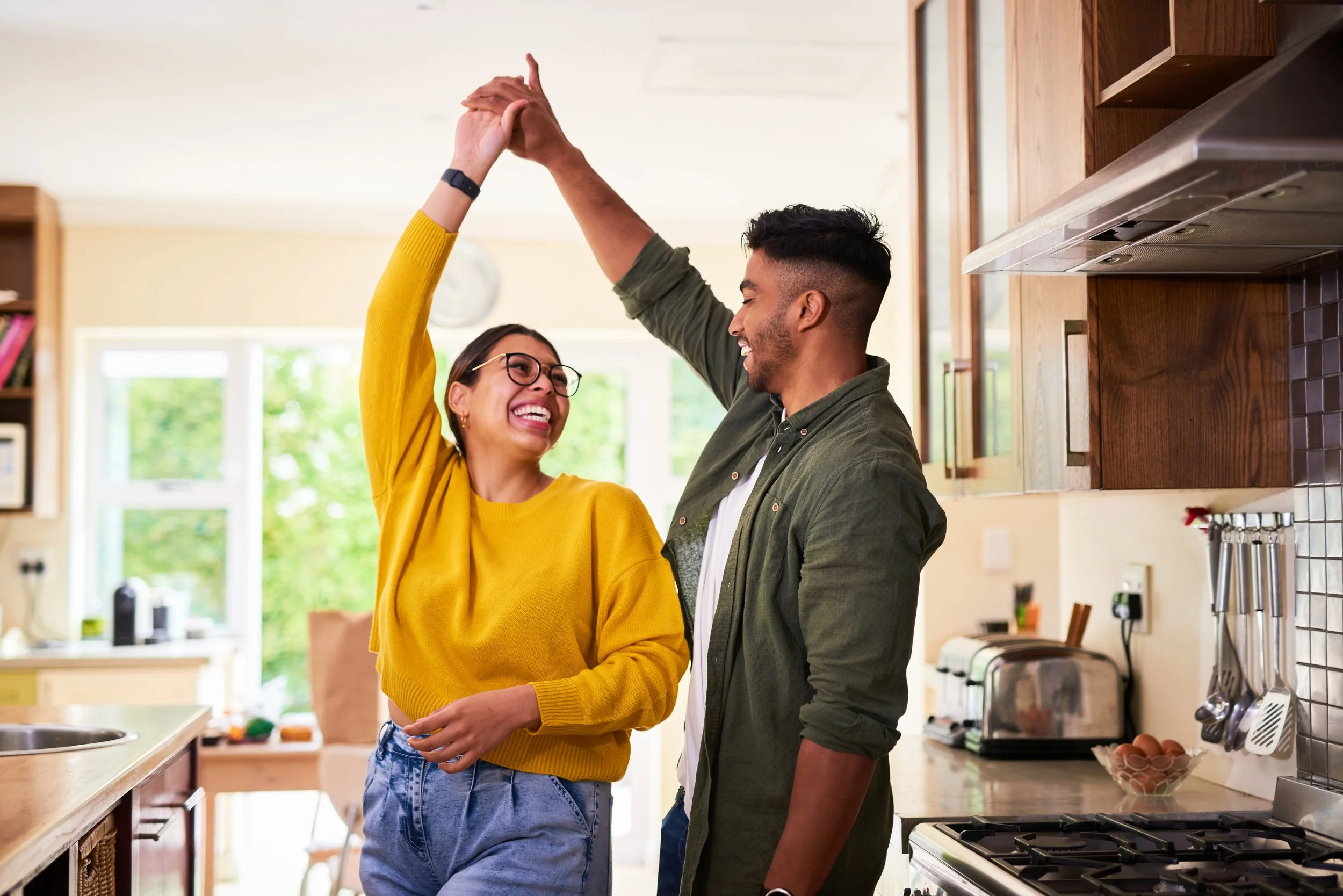 A young woman and man happily dance as he spins her in a bright kitchen, smiling at each other.