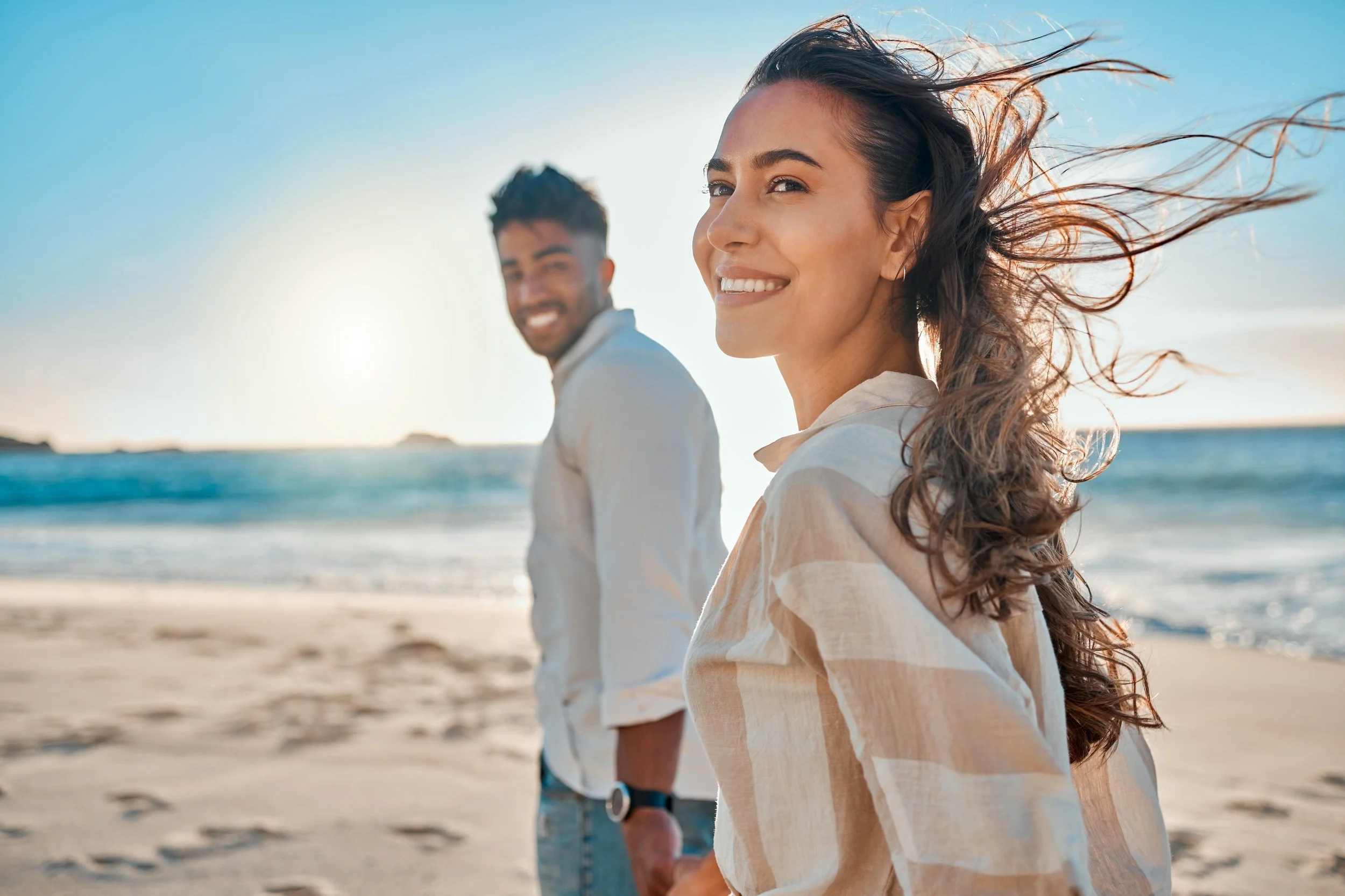 A woman with long hair blowing in the wind smiling at the camera on the beach, with a man standing behind her, near the ocean during sunset.