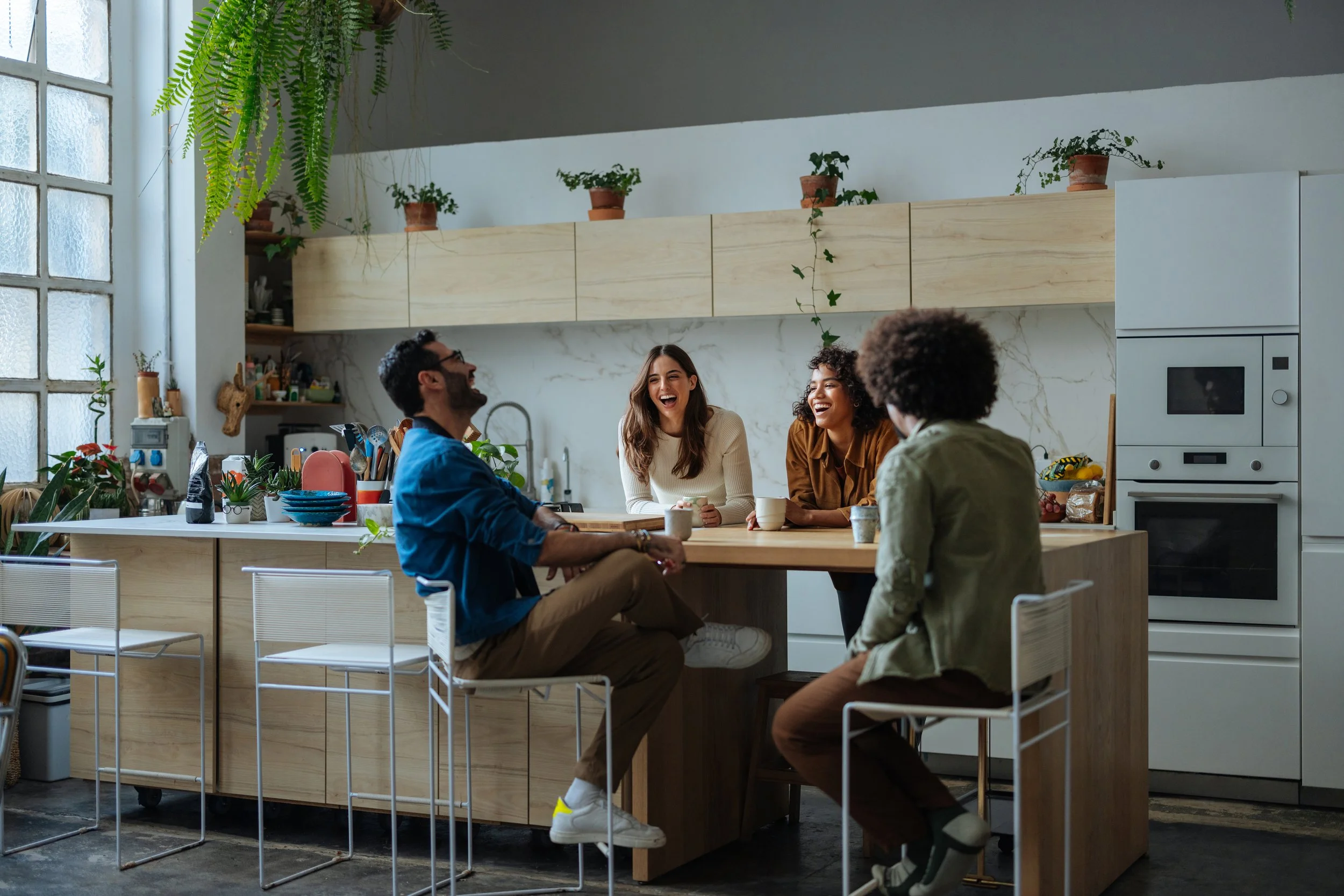 Four friends are sitting at a wooden kitchen island, laughing and enjoying company in a modern, well-lit kitchen decorated with potted plants and hanging greenery.