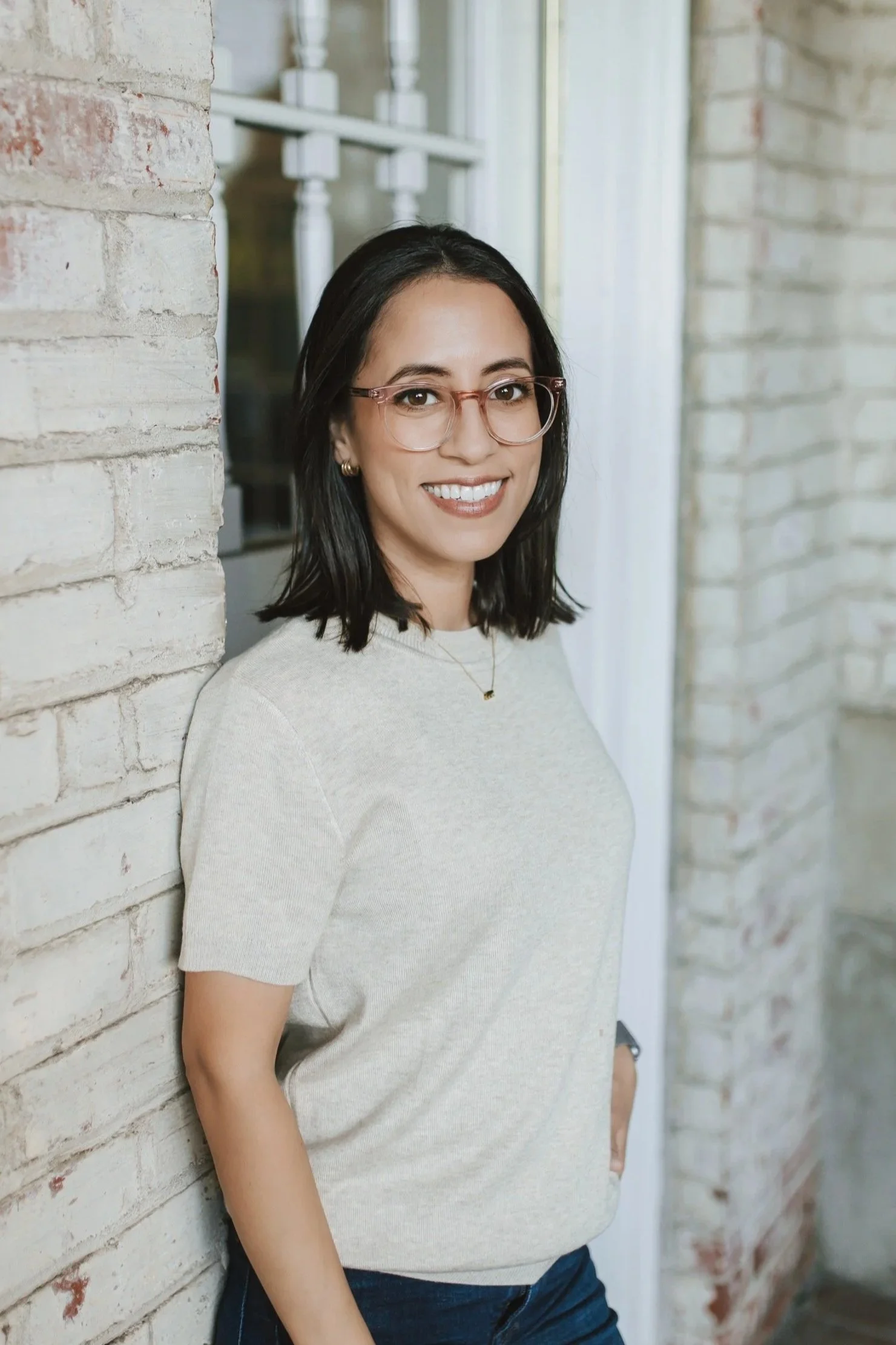A woman with shoulder-length dark hair, wearing glasses and a grey t-shirt, is standing near a brick wall, smiling at the camera.