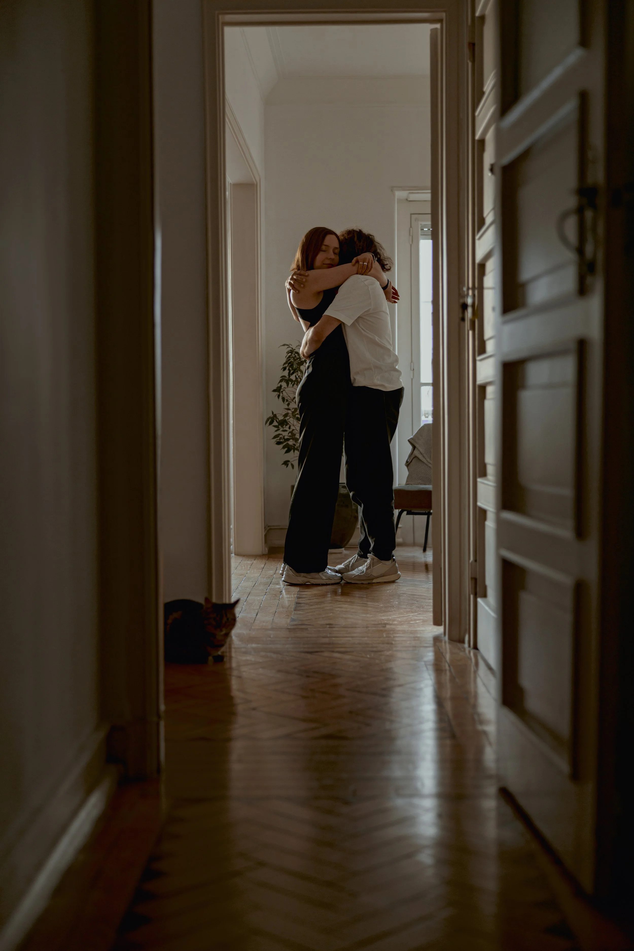 Two people embracing in a hallway, seen through a doorway, with a cat partially visible on the floor to the side.