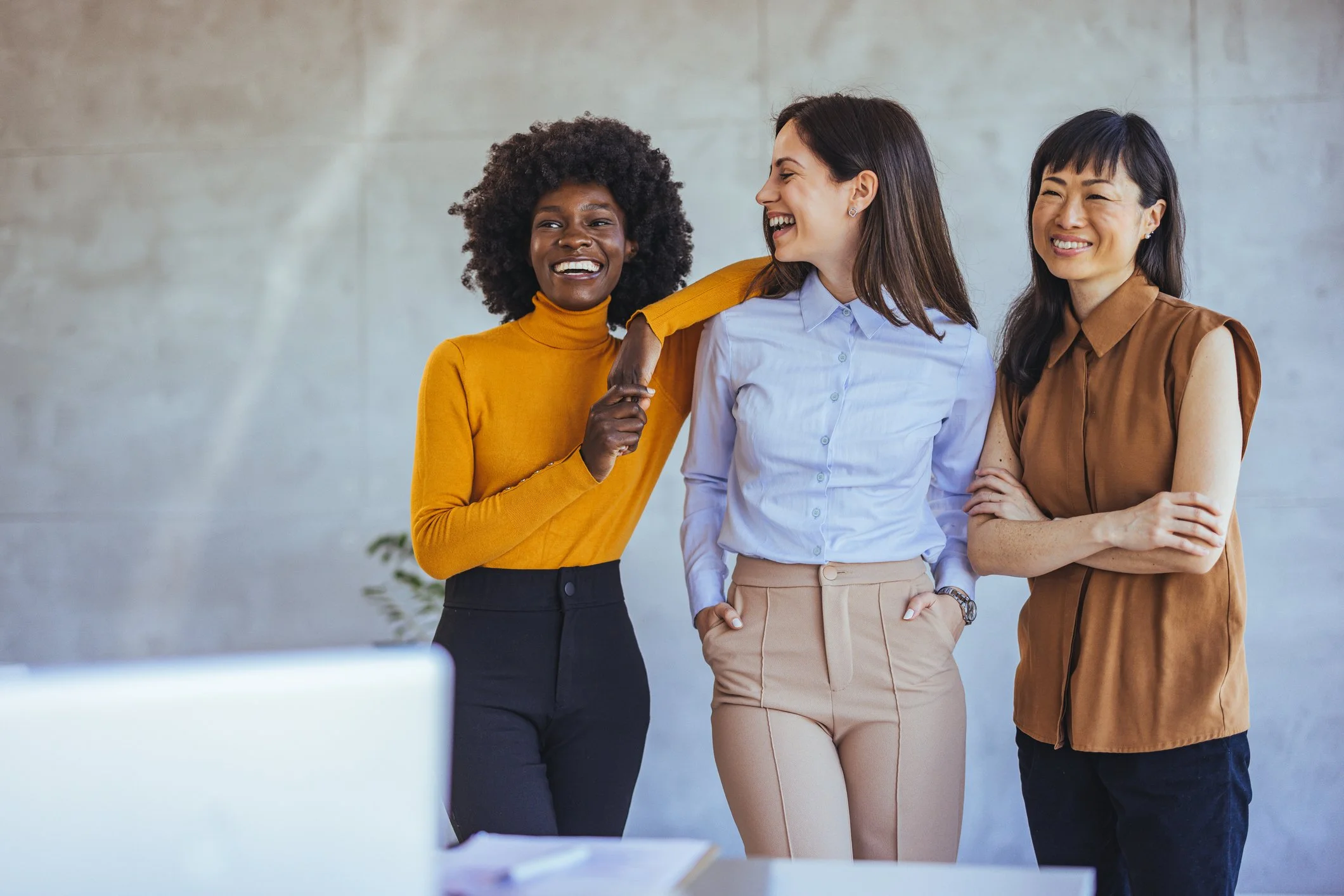 Three women smiling and standing close together in an office, with a desk and a computer in the foreground.