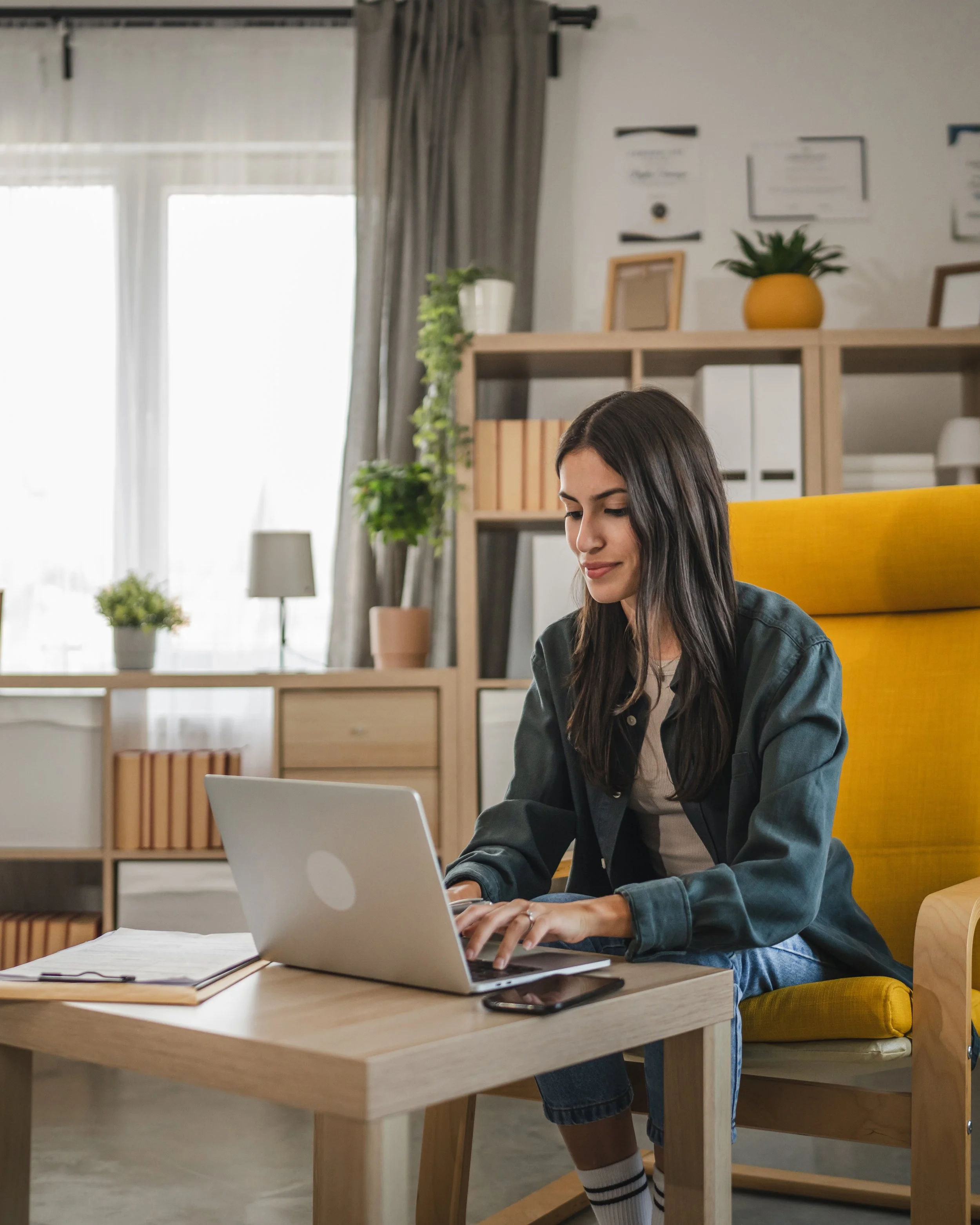 A young woman with long dark hair working on a laptop at a wooden desk in a modern, well-lit room. She is wearing casual clothes, including a yellow and grey striped sock visible on her leg, and sitting on a yellow chair. In the background, there are shelves with books, plants, and framed certificates or artwork on the wall.