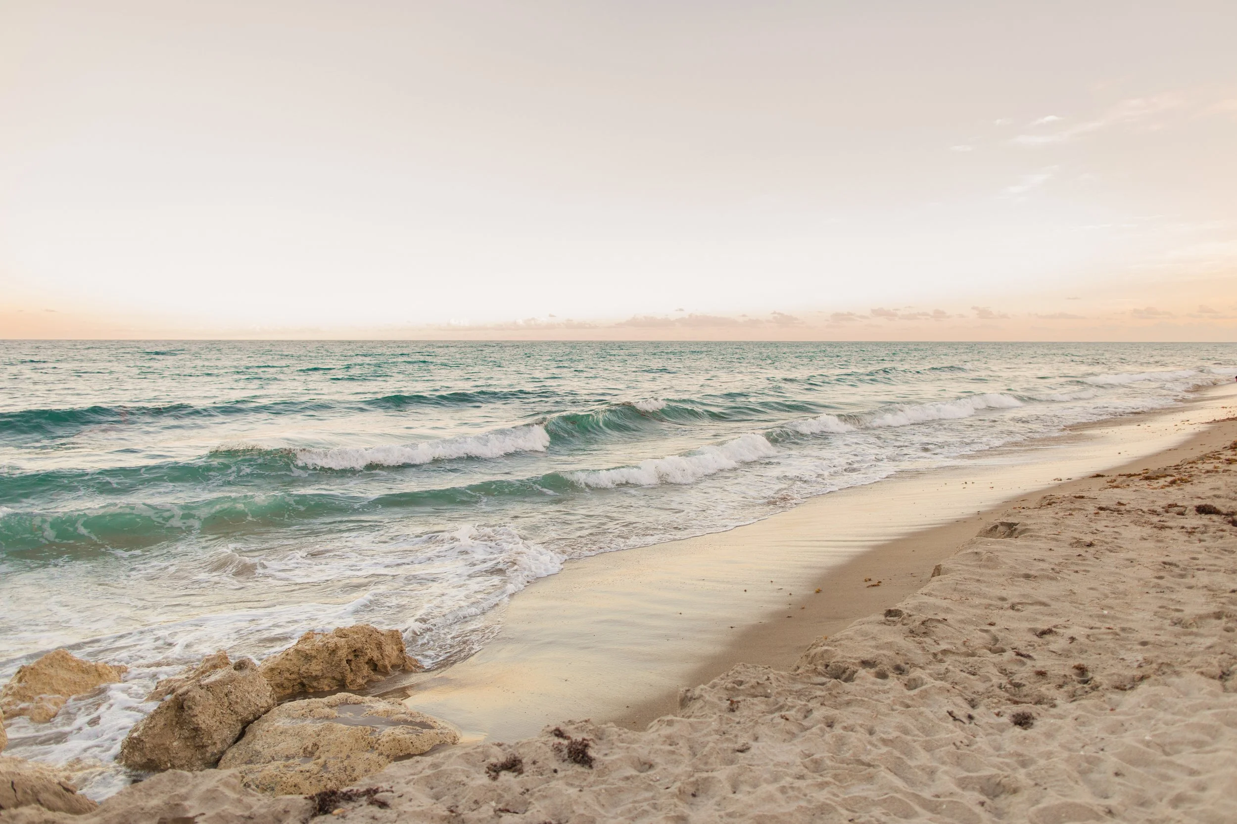 A sandy beach with rocks in the foreground and gentle ocean waves reaching the shore. The sky is soft and pastel-colored, indicating early morning or late evening.