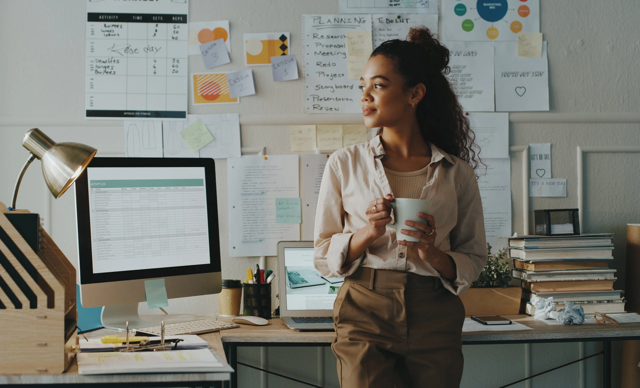 A woman standing in an office, holding a mug, surrounded by books, a computer, and a cluttered desk. The wall behind her is covered with notes, charts, and papers related to planning and design.
