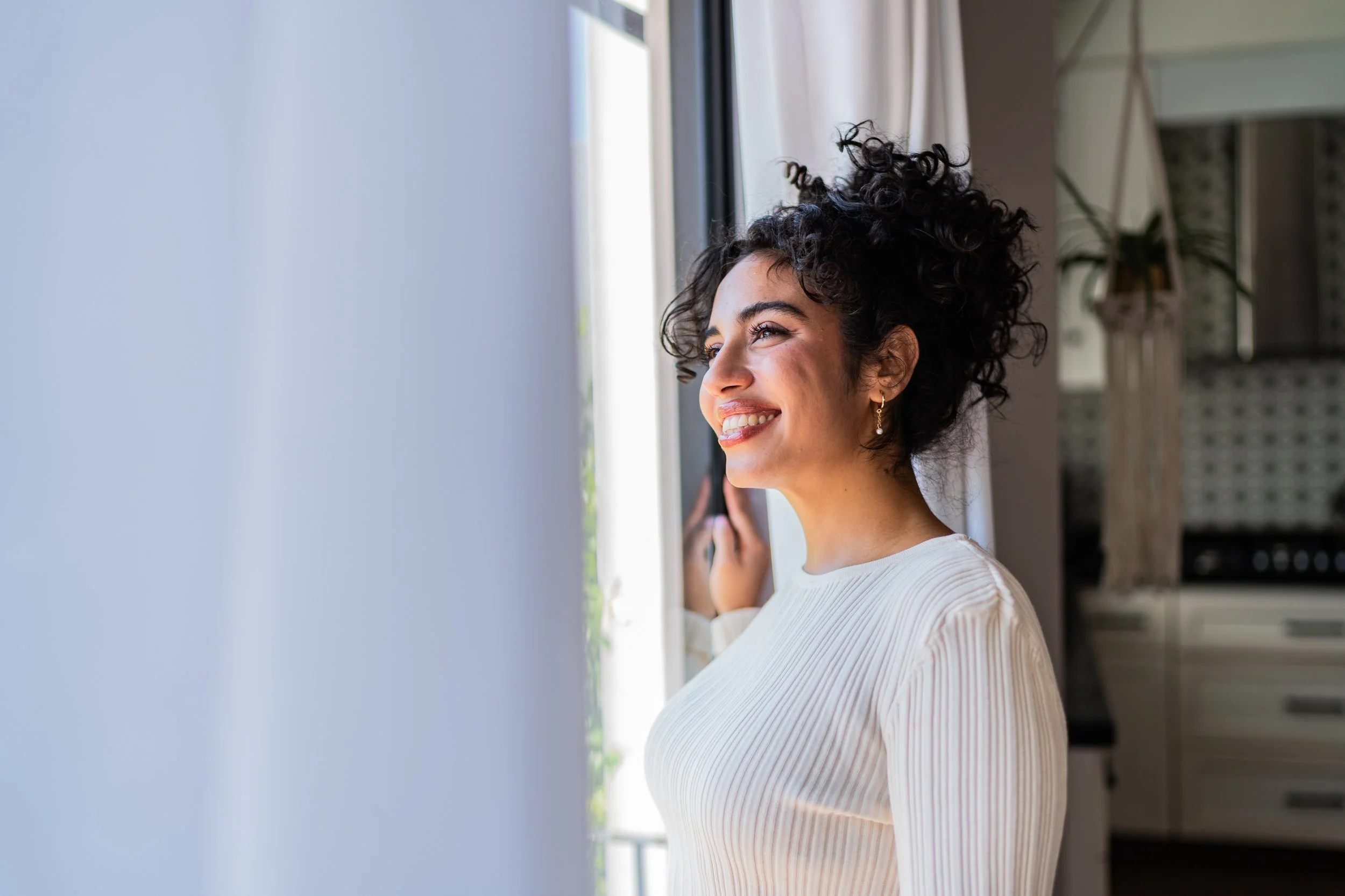 Close-up of a woman with curly dark hair smiling while looking out a window in a bright, modern room.
