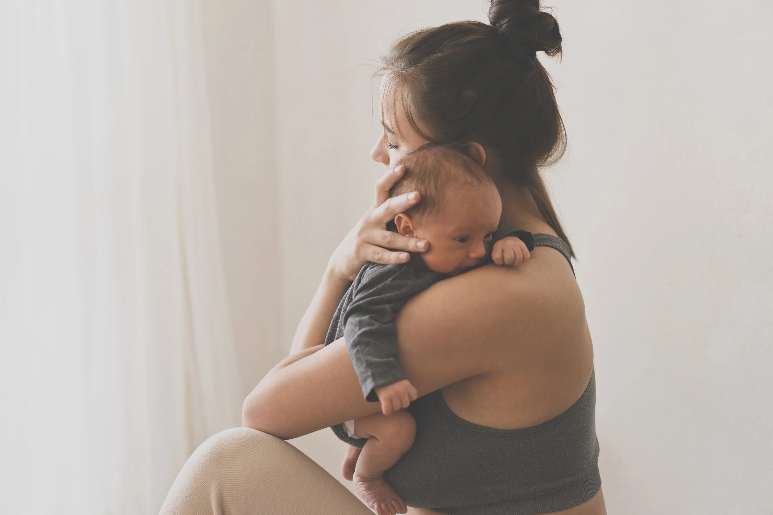 A woman with brown hair in a bun holds a sleeping baby with curly hair close to her chest, standing against a plain white wall.