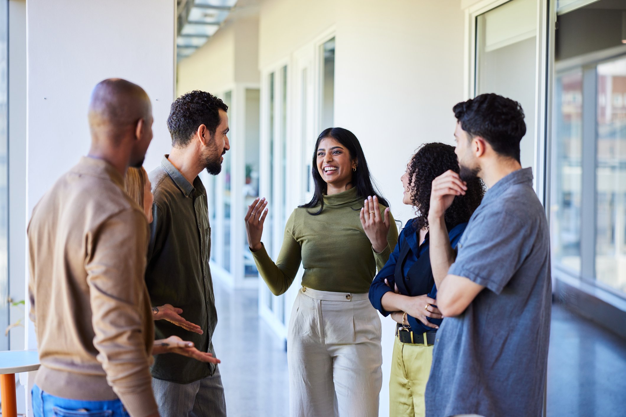 Group of five diverse adults standing in a modern office, engaged in conversation. The woman in the center is smiling and talking, while others listen.