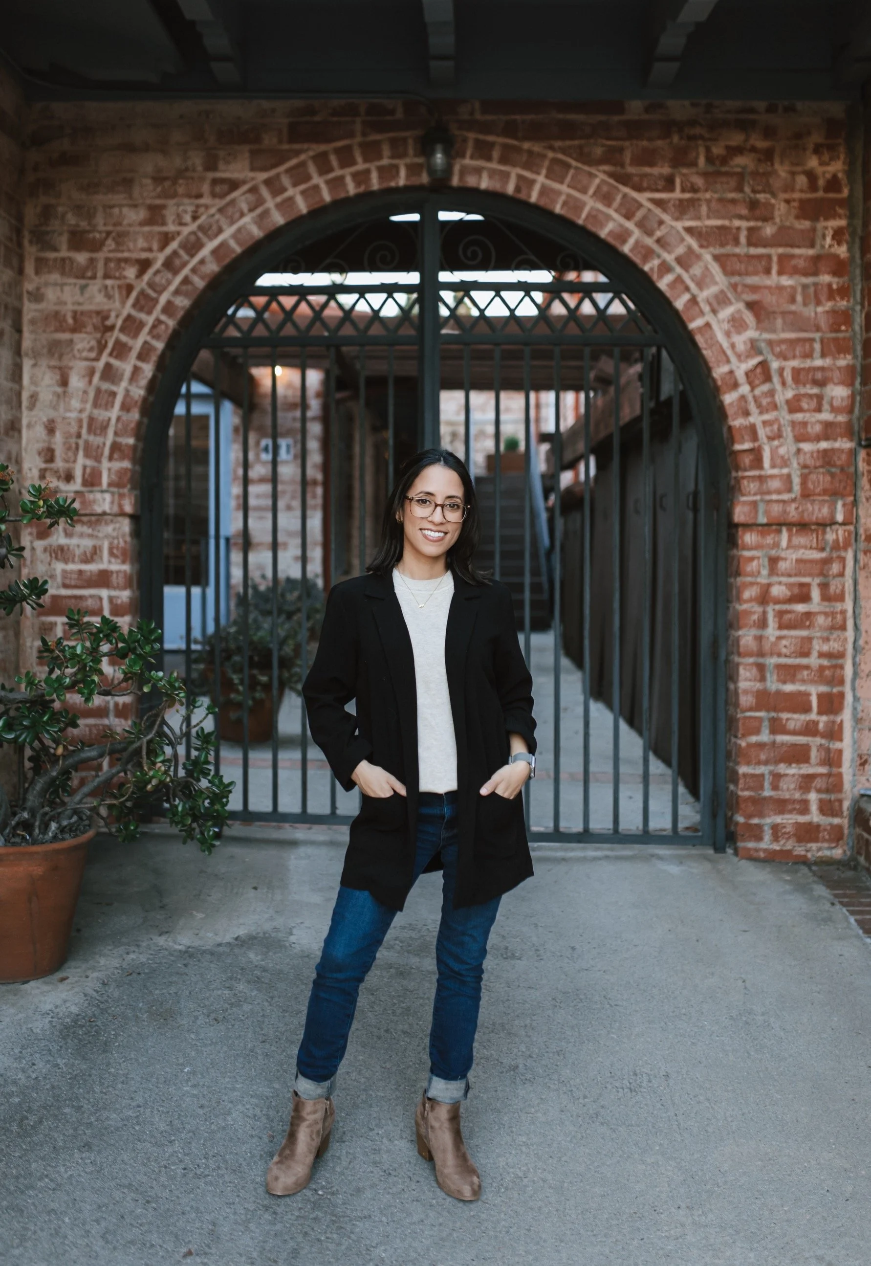 Woman standing outdoors in front of a brick archway with black gates, wearing glasses, a black coat, gray sweater, jeans, and tan boots, smiling.
