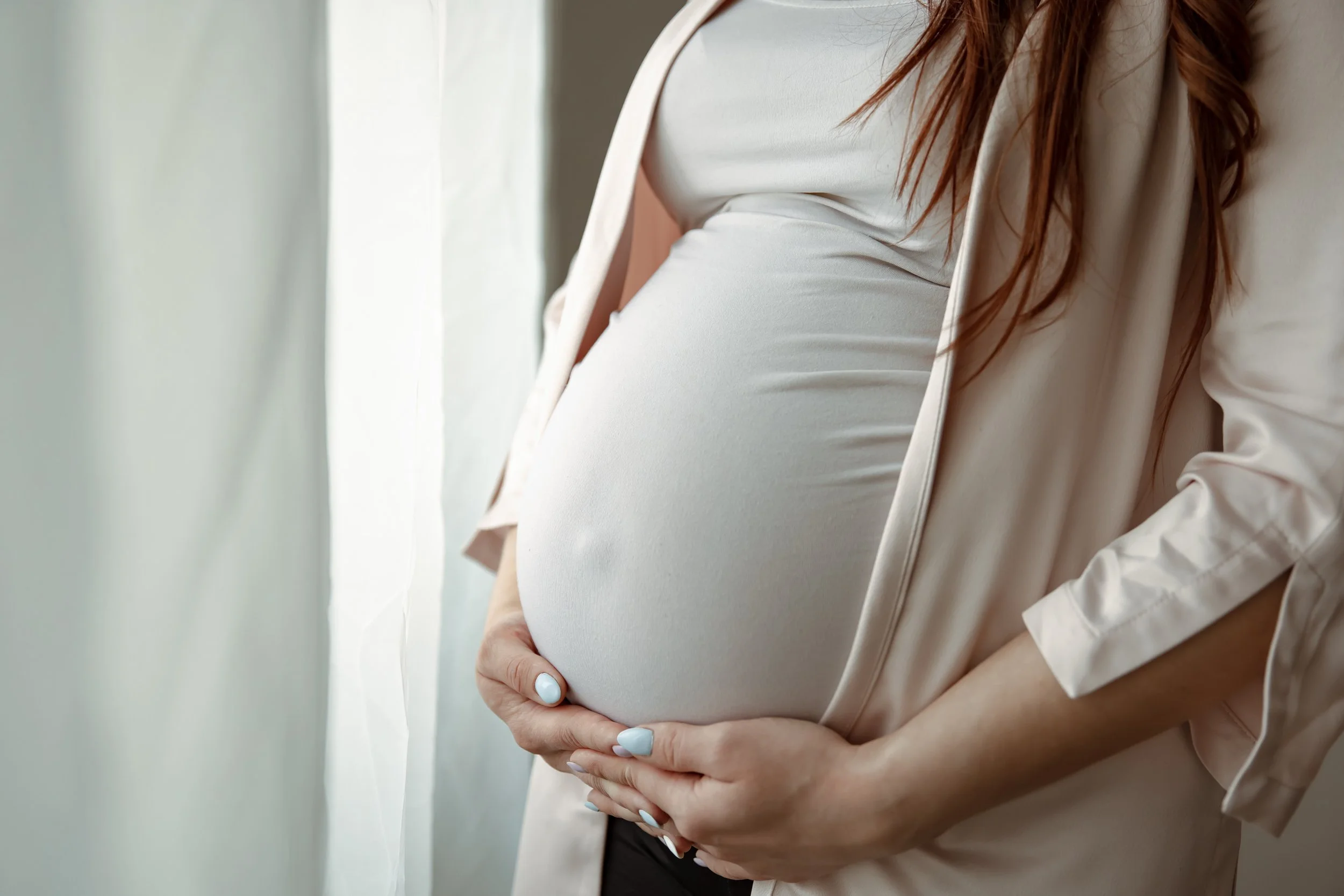 A pregnant woman with long red hair wearing a white top and beige blazer gently holding her belly, standing near a window with soft natural light.