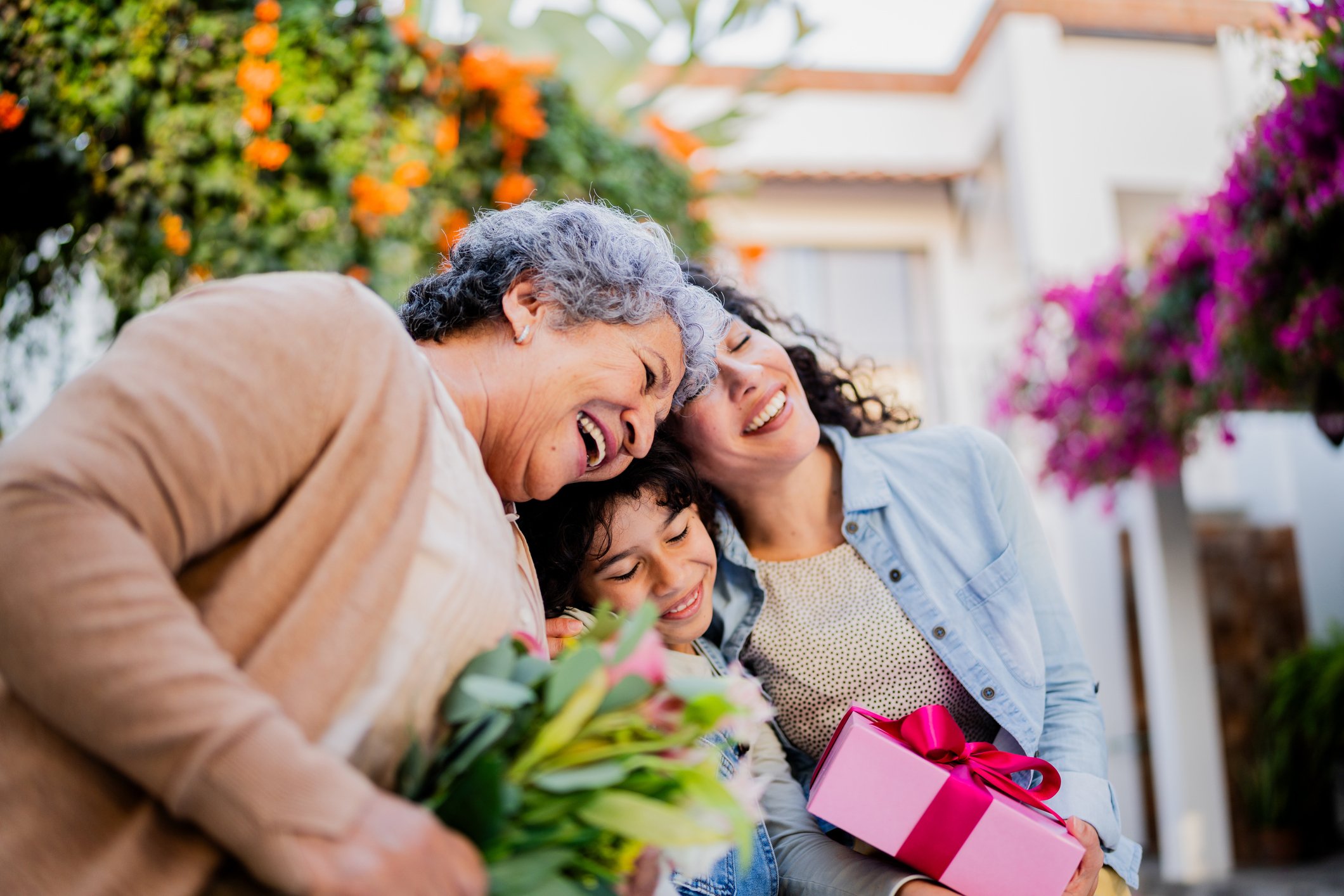 Grandmother, mother, and child smile, with the mother holding a pink and white gift box with a pink ribbon, surrounded by colorful flowers in a garden.