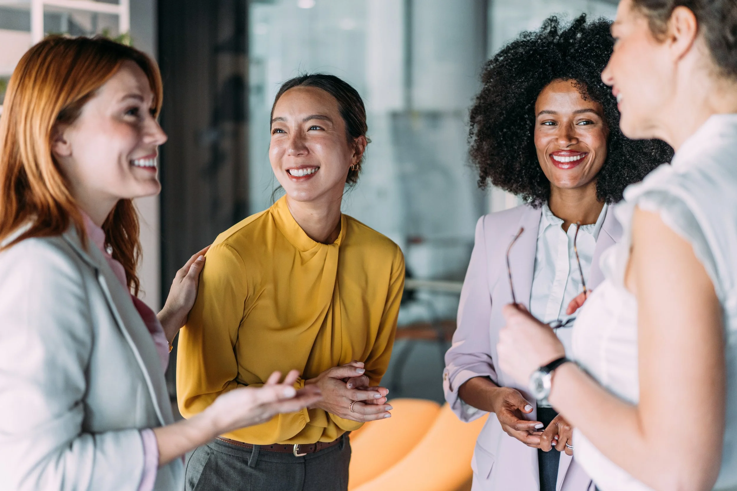 A diverse group of four women engaged in conversation indoors, smiling and appearing to enjoy each other's company.