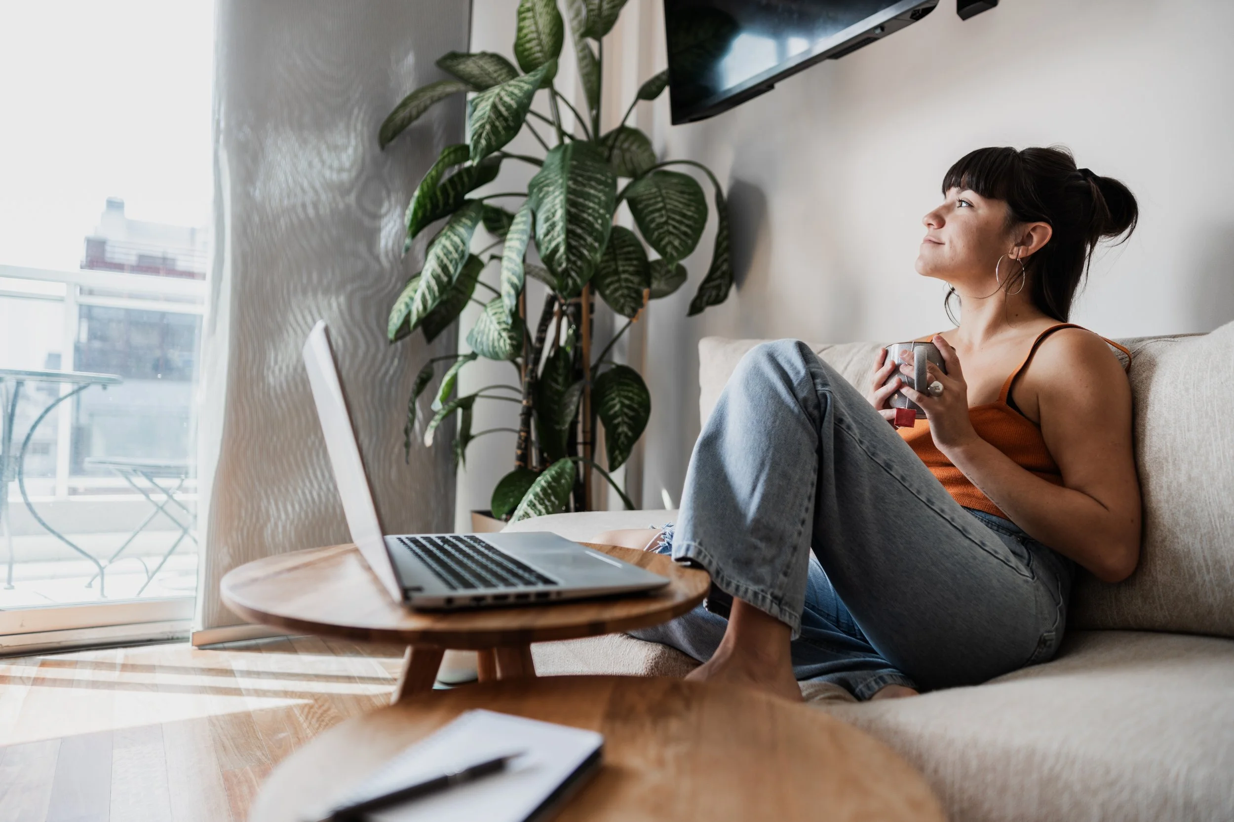 A woman sitting on a beige couch with her legs crossed, holding a mug, looking out a window with sheer curtains. A laptop is on a wooden coffee table in front of her, and a large green plant is beside her. A flat-screen TV is mounted on the wall above her.