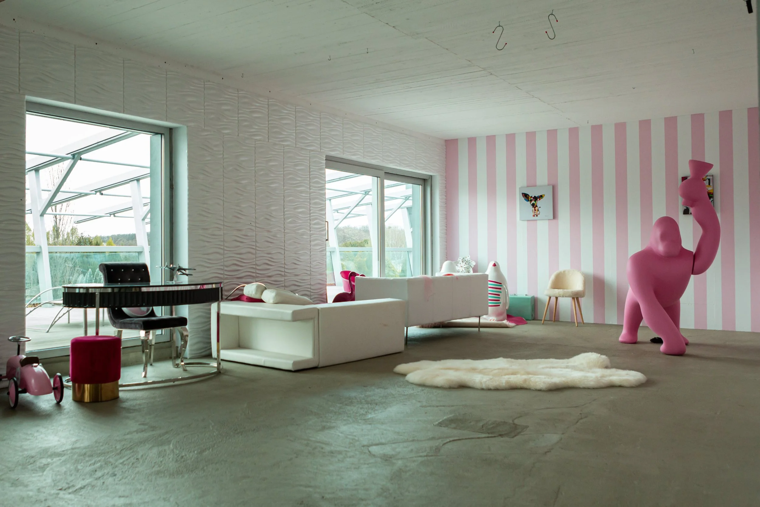A modern living room with pink and white striped wall, white textured wall, and large windows showing outdoor scenery. The room features playful decor including a large pink sculpture, a fluffy cream rug, and a white sofa. There are various chairs, t