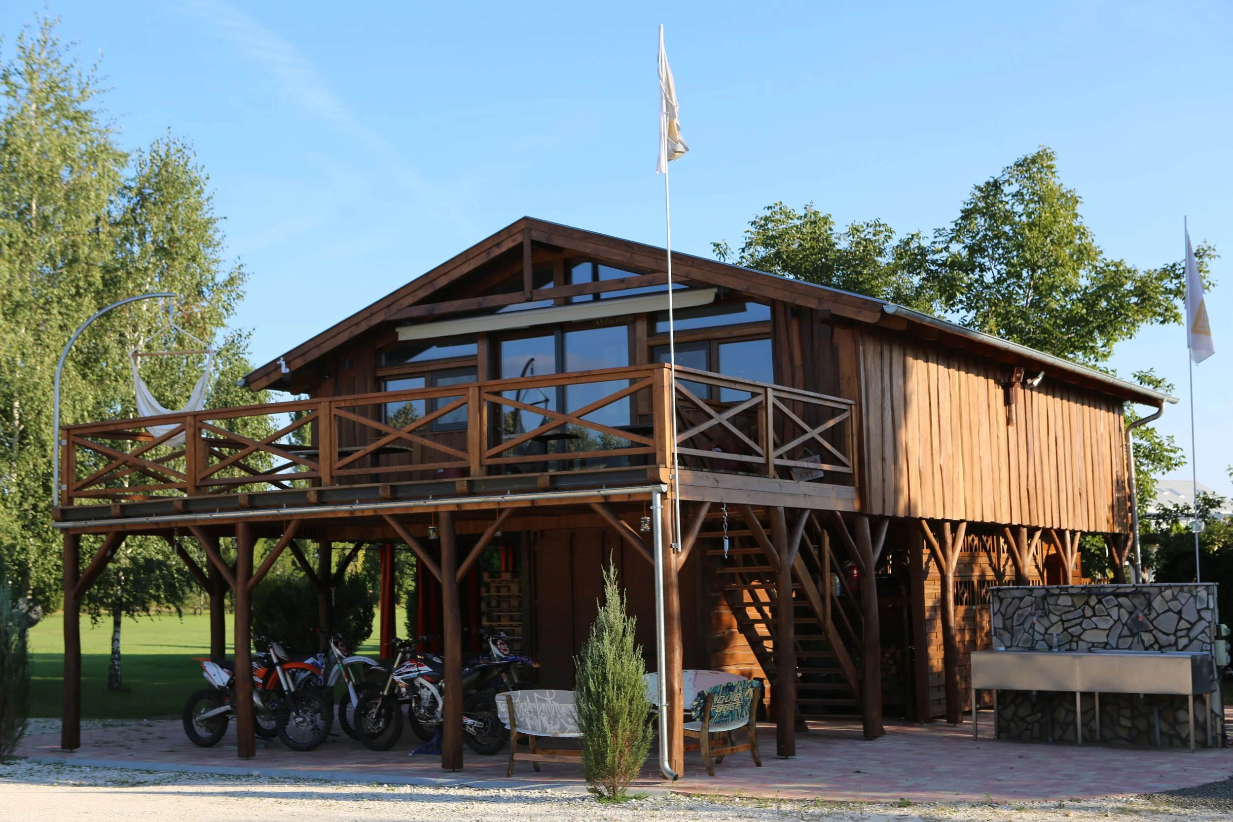 Two-story wooden house with a large outdoor balcony, flags on poles, motorcycles parked underneath, and a small seating area with chairs and a small tree in front, set in a sunny outdoor environment with green trees and a clear blue sky.