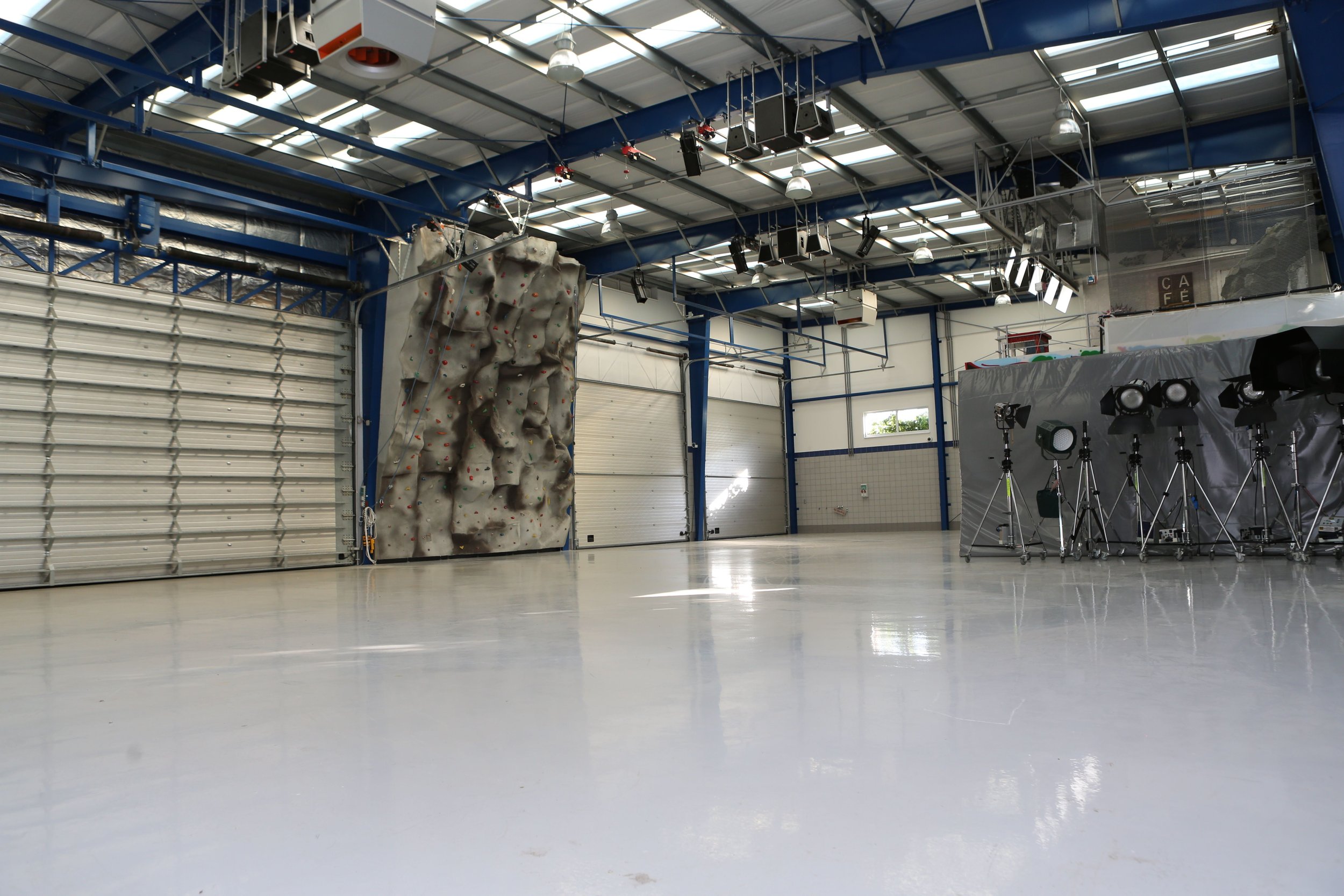 Empty indoor sports facility with a climbing wall, movable garage doors, and photography lighting equipment.