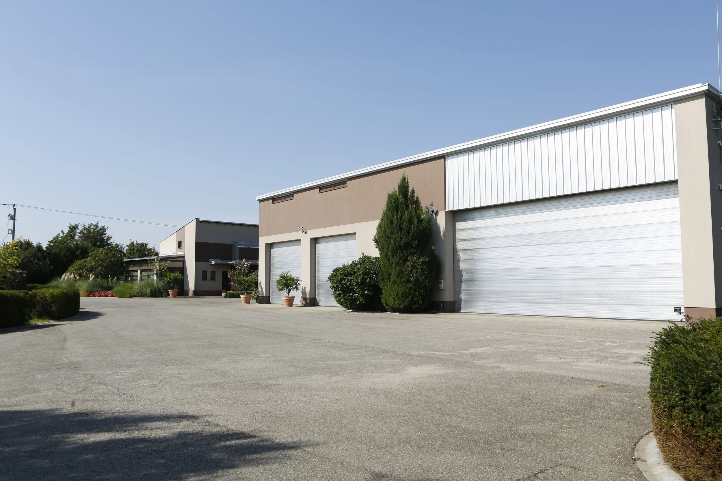 Commercial building with three large white roll-up garage doors, surrounded by potted plants and greenery, under a clear blue sky.