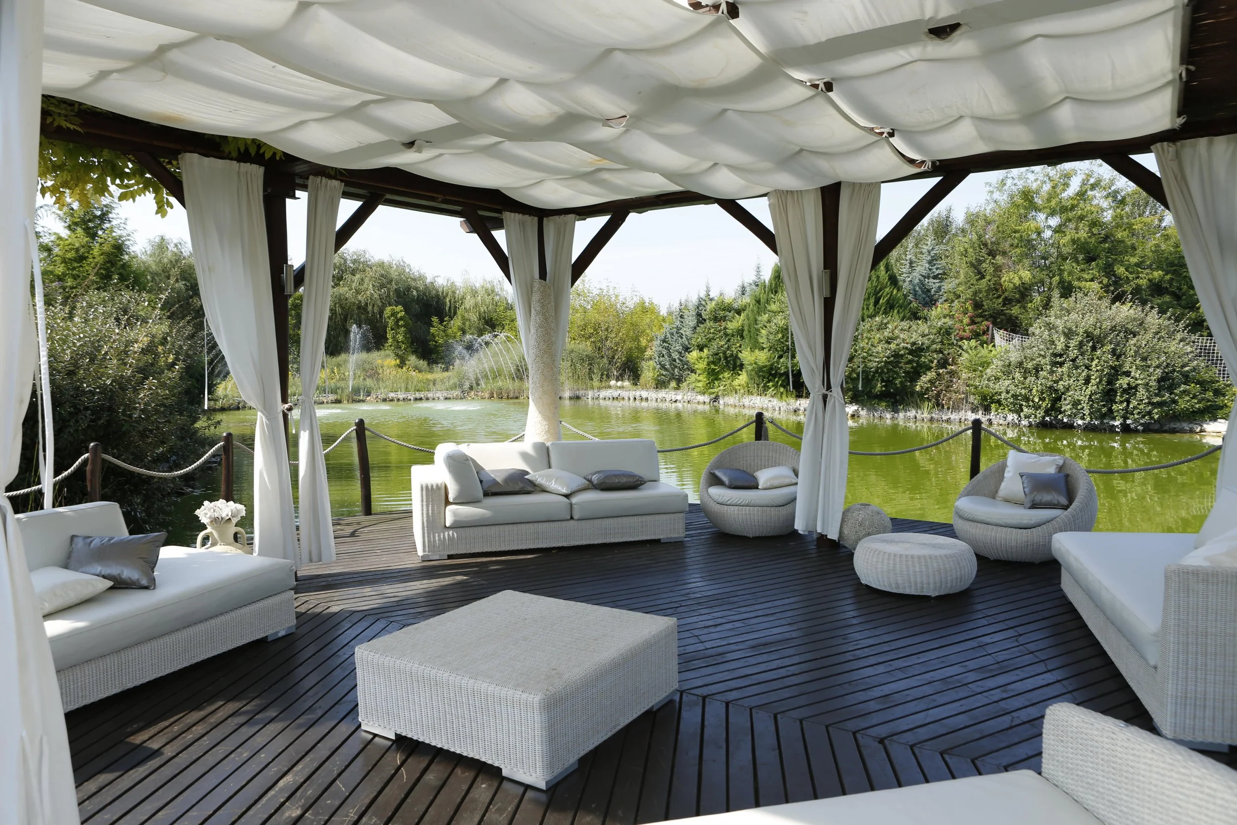 Outdoor gazebo with white wicker and fabric furniture overlooking a pond, surrounded by lush green trees.