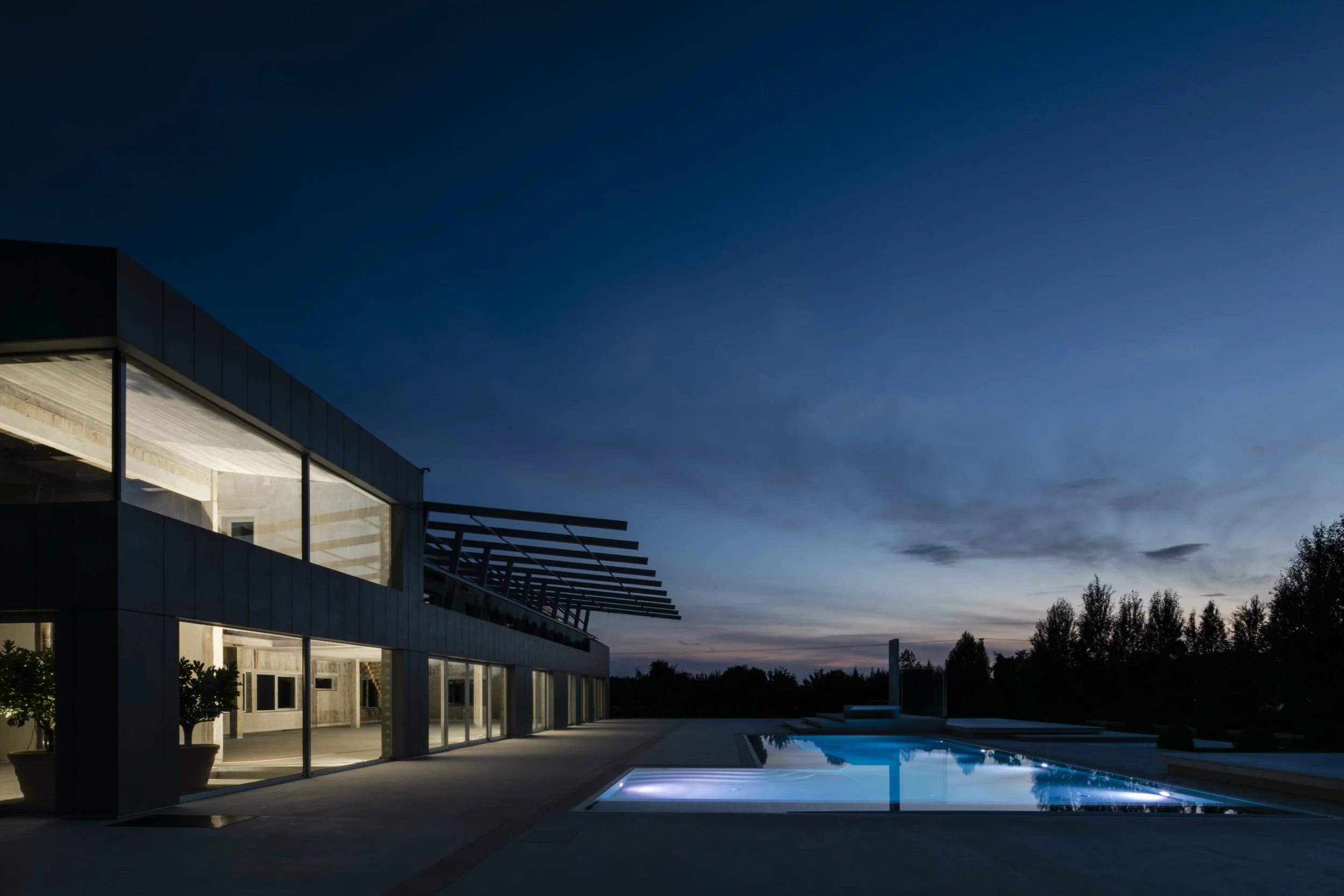 Modern house with a large glass exterior, outdoor pool, and patio area, at dusk with a darkening sky and trees in the background.