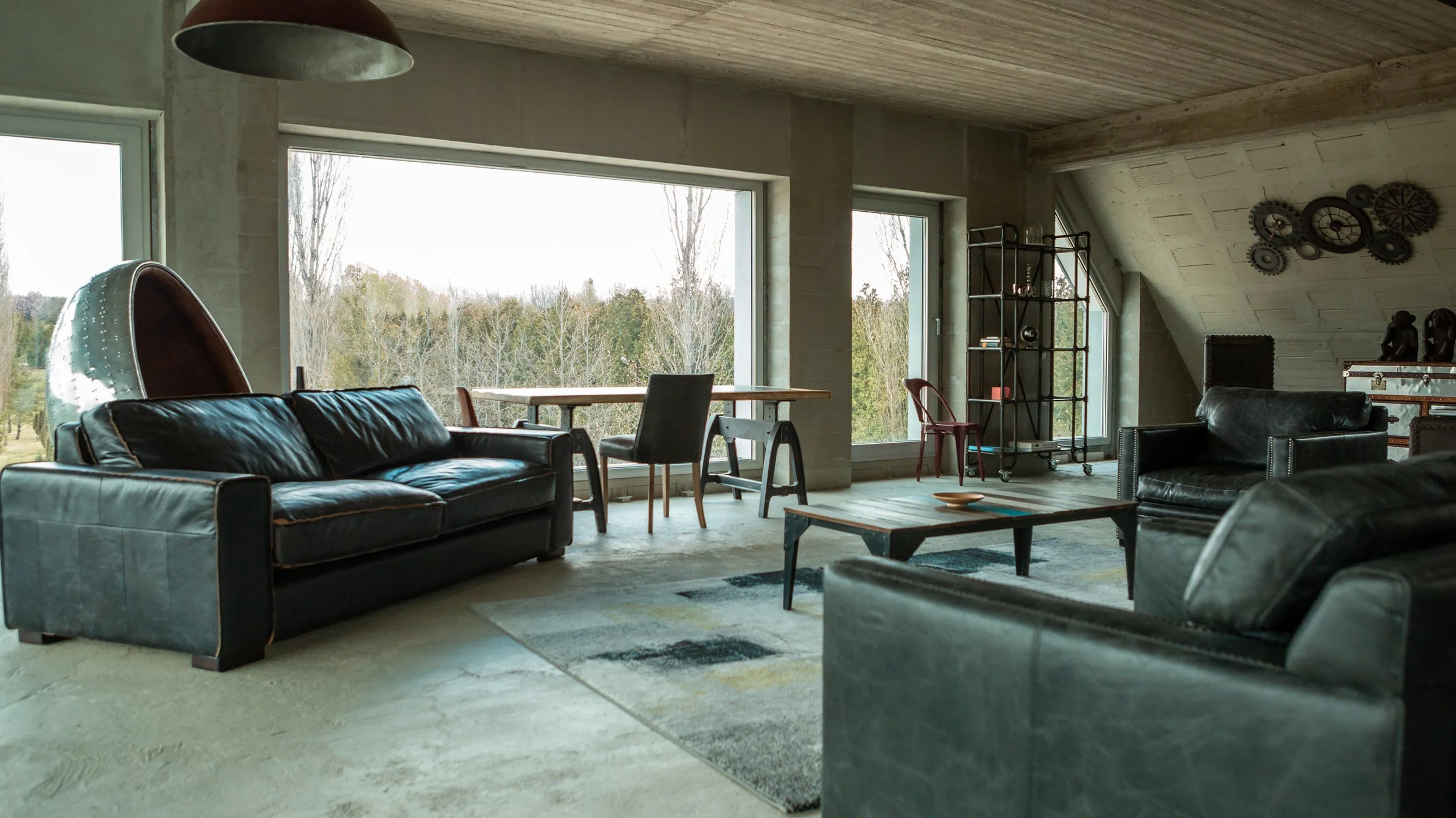 Modern living room with large windows, black leather sofas, a wooden table, colorful area rug, and decorative wall gears.