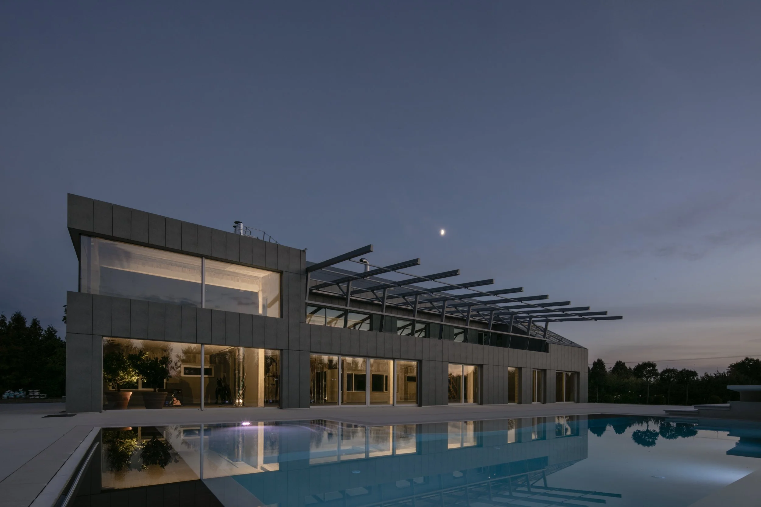 Modern house with large glass windows and a rooftop structure, reflected in a swimming pool at dusk with a visible moon in the sky.