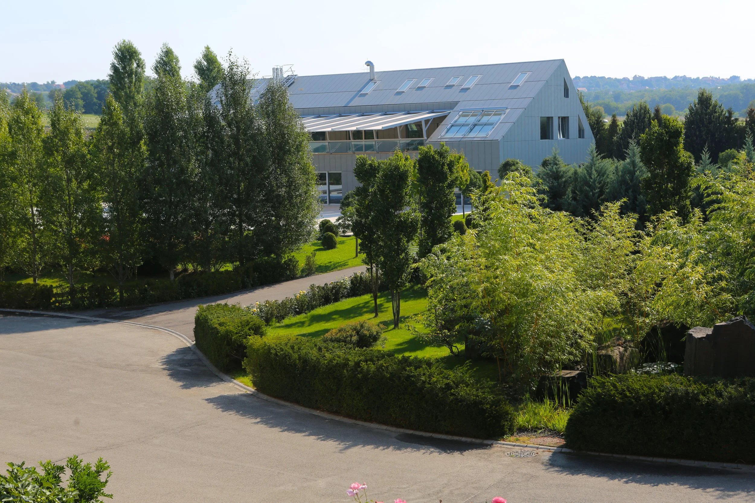 Modern building with a sloped roof, surrounded by lush green trees and a landscaped garden, with a paved driveway in front.