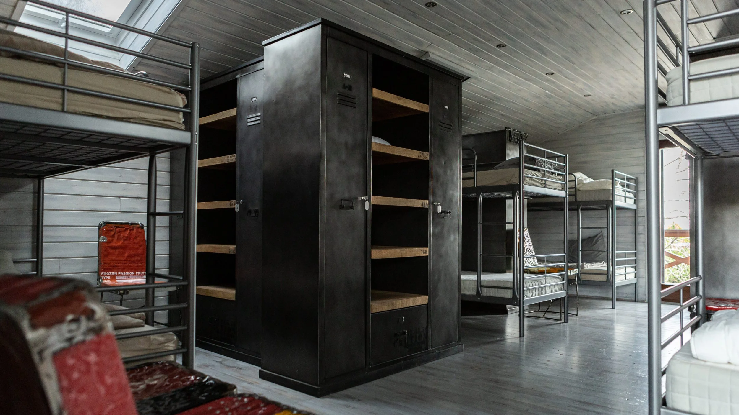 A room with multiple bunk beds and black lockers, with wooden shelves inside some lockers, and a gray wooden ceiling and floor.